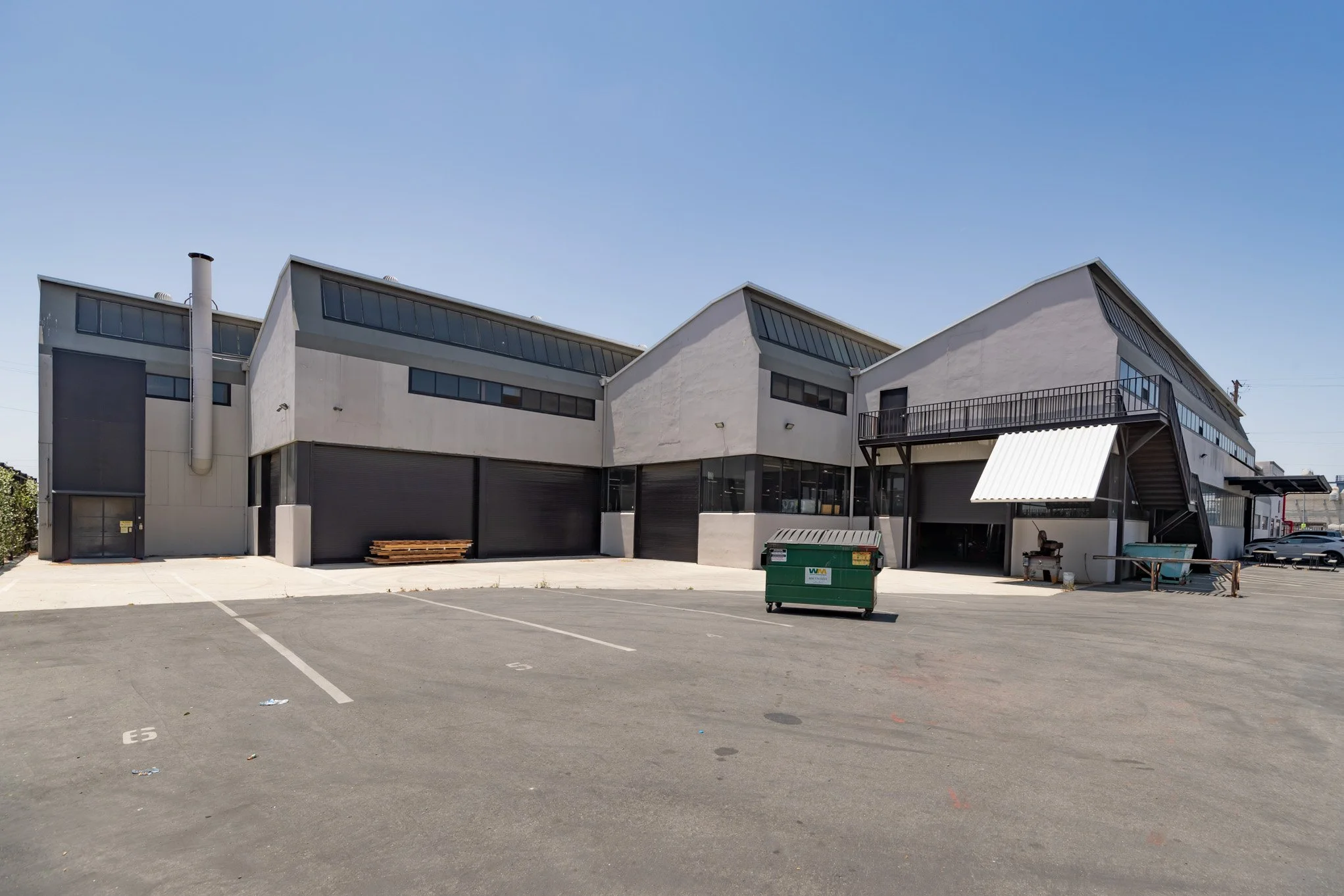 A commercial building with multiple levels, large windows, and black roller doors, with a parking lot in the foreground and a blue sky above.