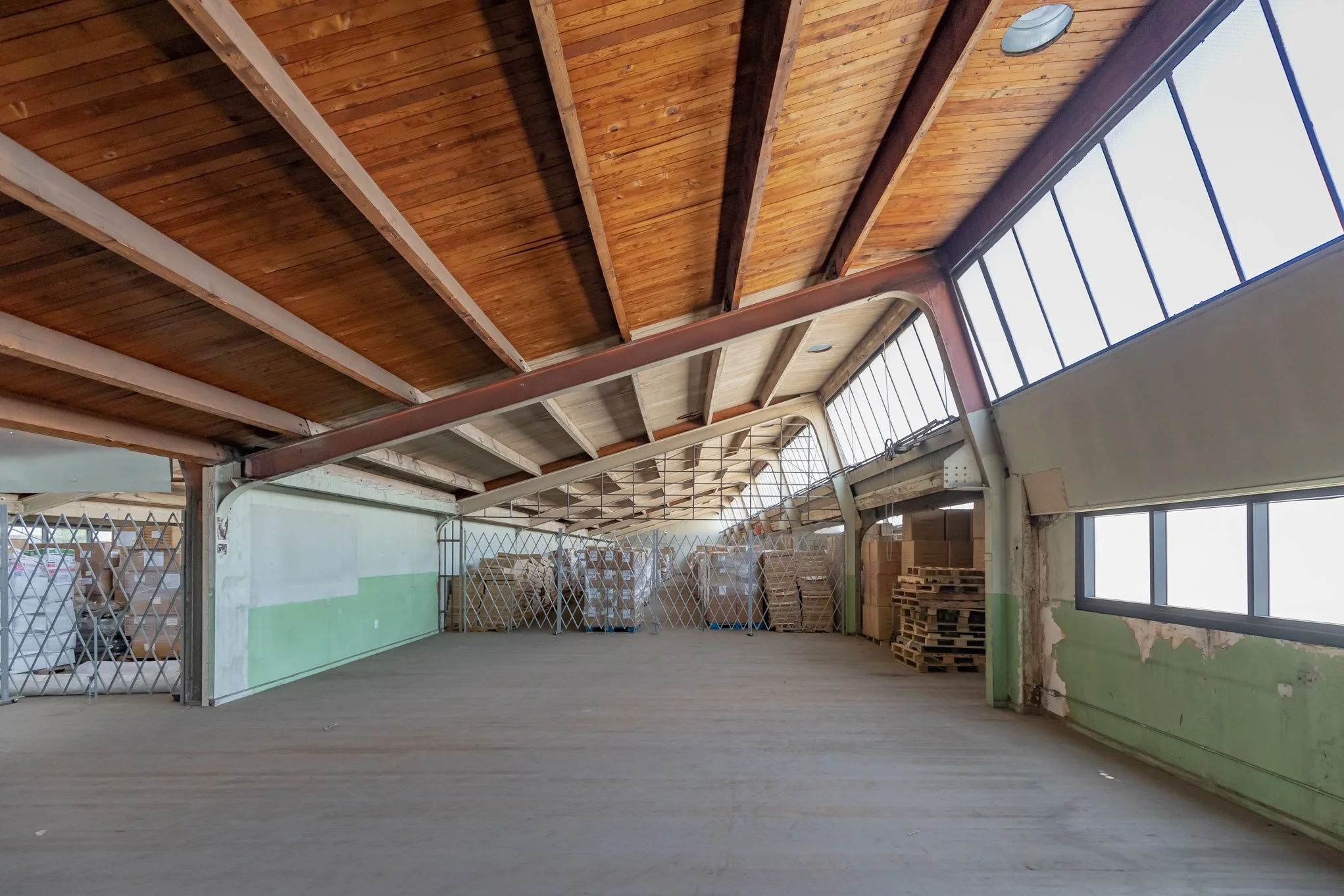 Empty warehouse with wooden ceiling, large windows, stacks of pallets, and stored boxes behind a retractable security gate.