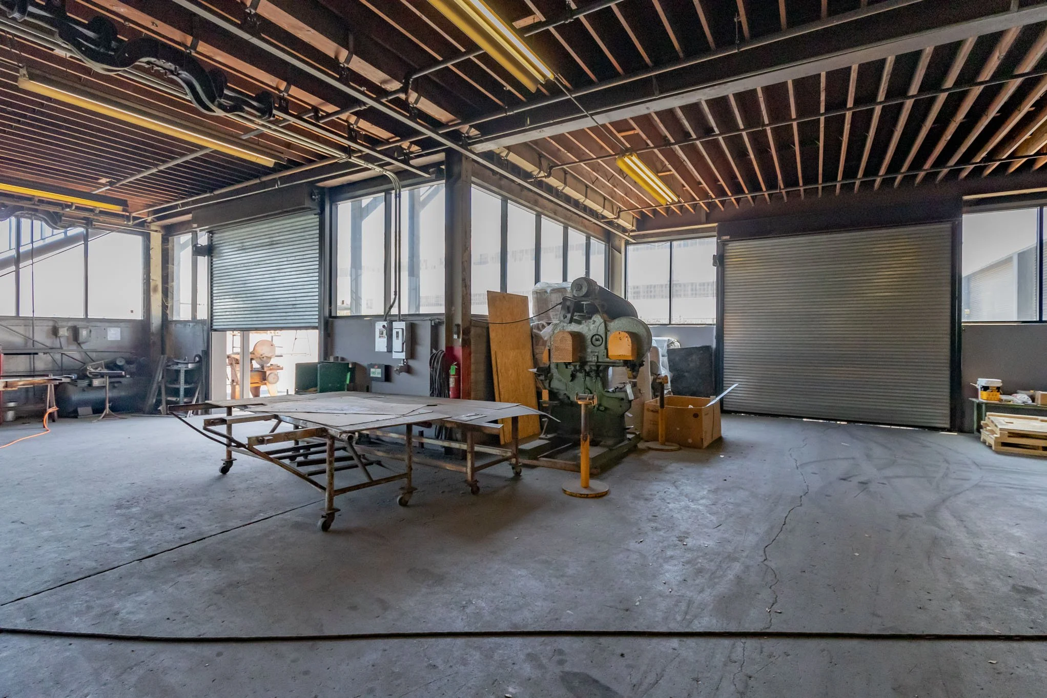 Empty industrial workshop with large windows, metal rolling door, and scattered equipment including a table and machinery.