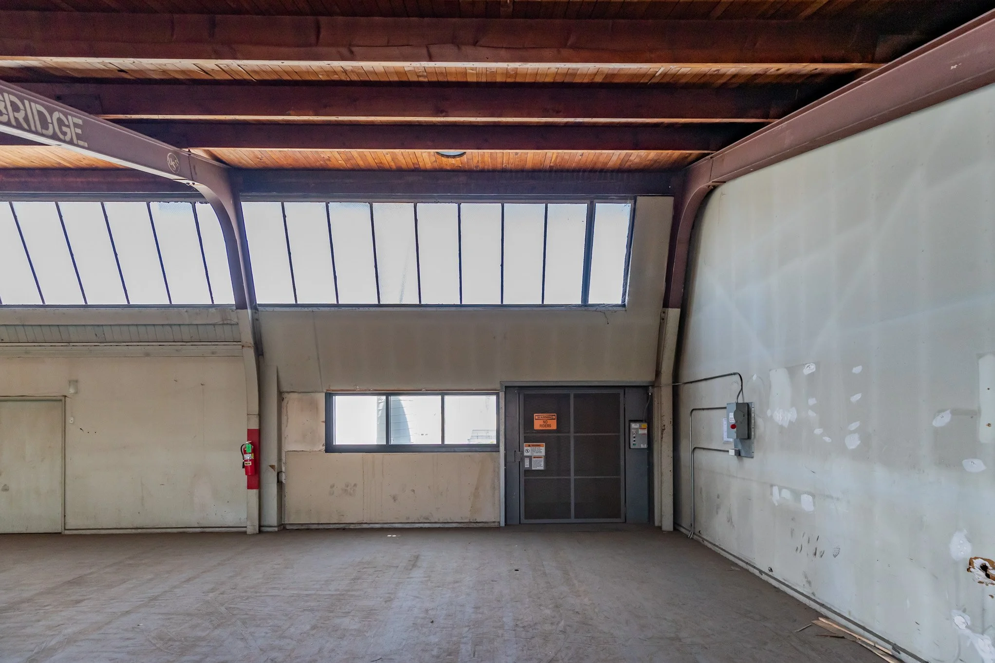 Empty industrial or warehouse space with concrete floor, large window, metal door, and fire extinguisher on wall.
