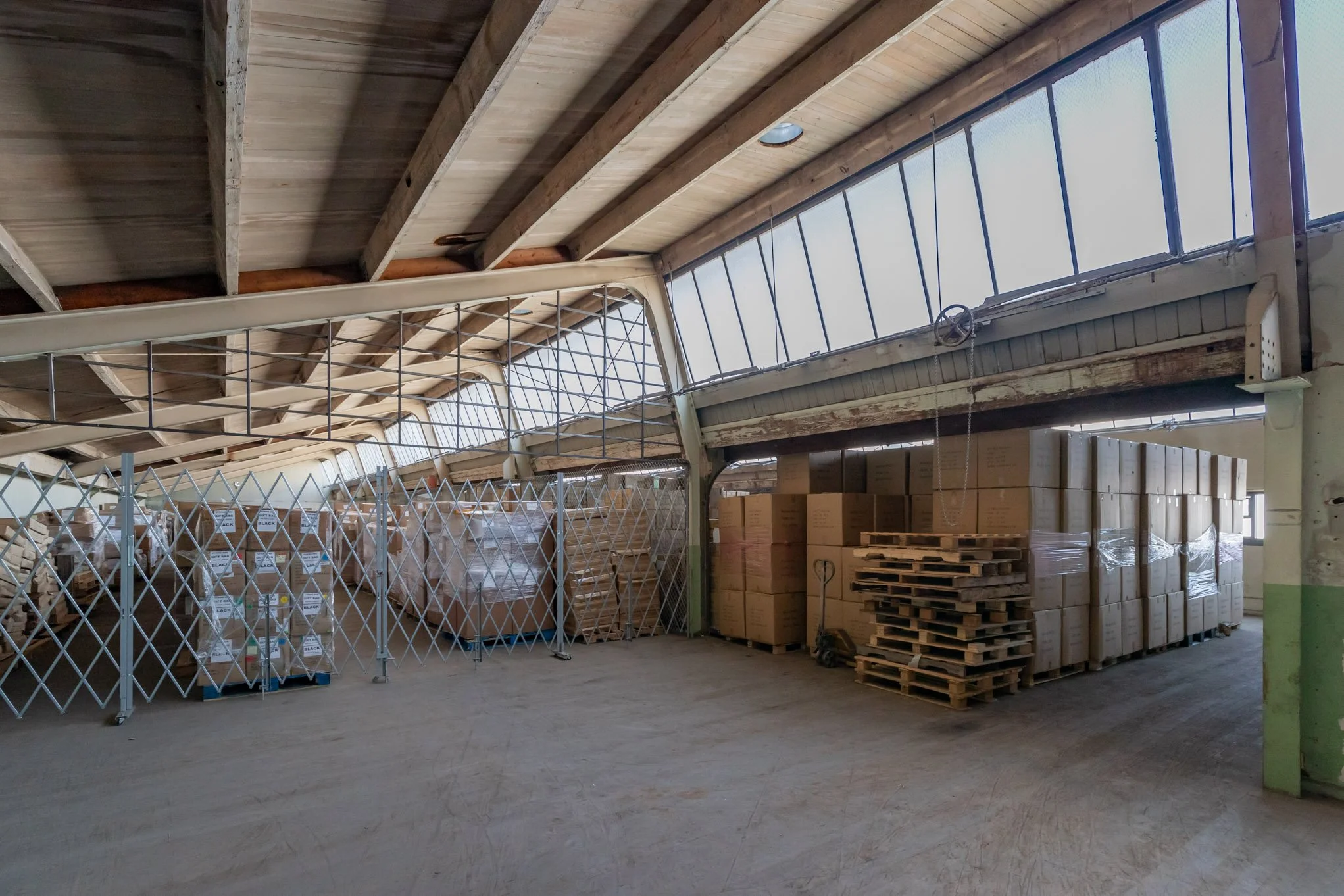 Inside an industrial warehouse with stacked pallets of boxes and wood, a partially open rotating warehouse door, and a wooden ceiling with skylights.