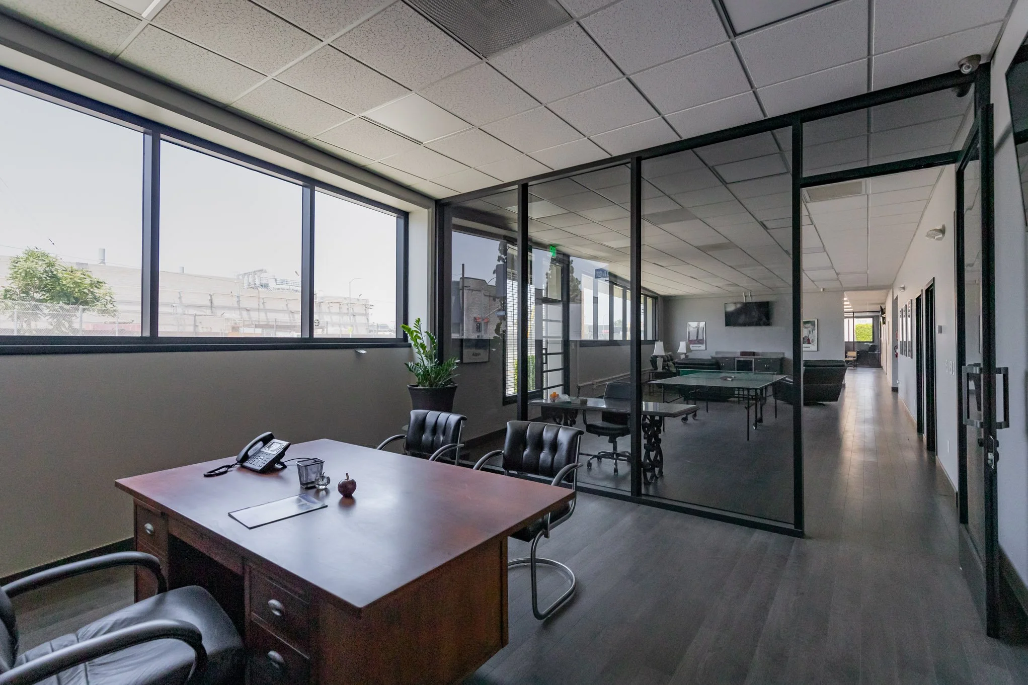 Modern office with a wooden desk, black chairs, a landline phone, and a small decorative item in the foreground. Behind a glass partition, there is a seating area with couches, a ping pong table, and office furniture. Large windows allow natural ligh