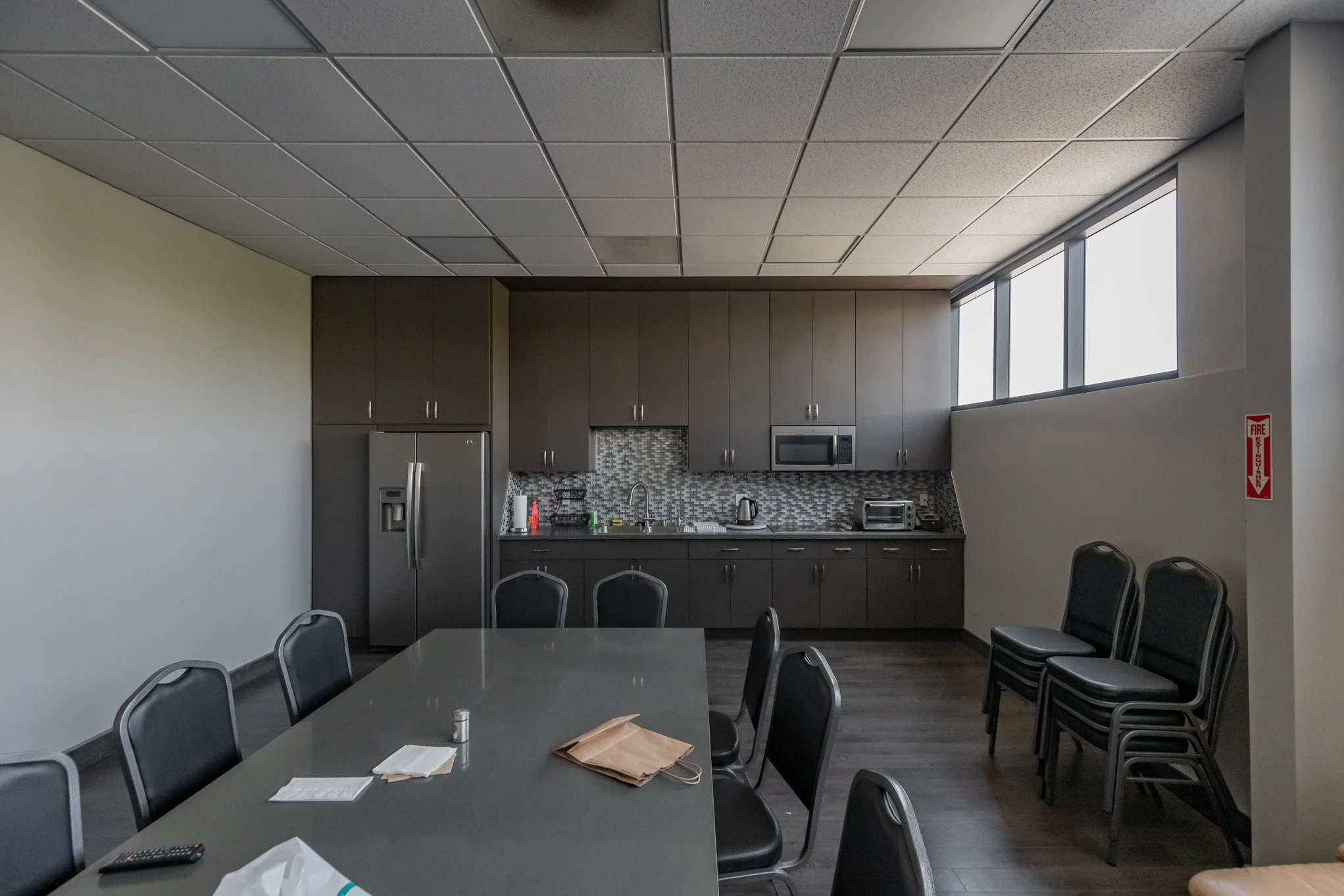 Empty conference room with a long table, black chairs, kitchenette with gray cabinets, microwave, toaster, and a window high on the wall.