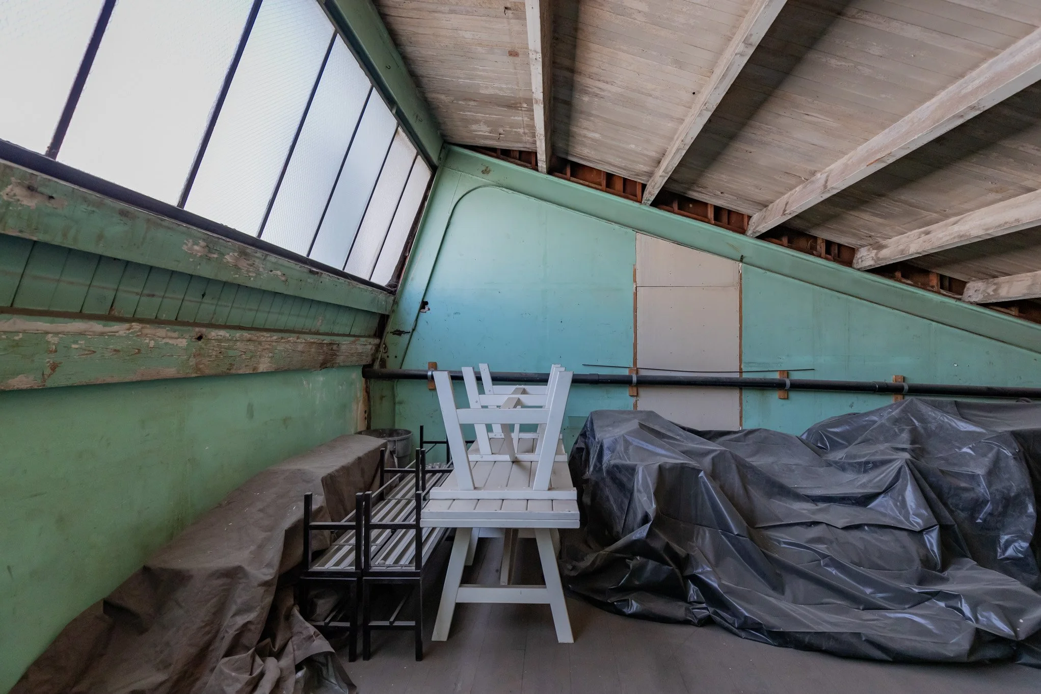 Unfinished attic with plastic-covered items, a white wooden chair, and a small black metal frame, with green and blue walls and exposed wooden beams on the ceiling.