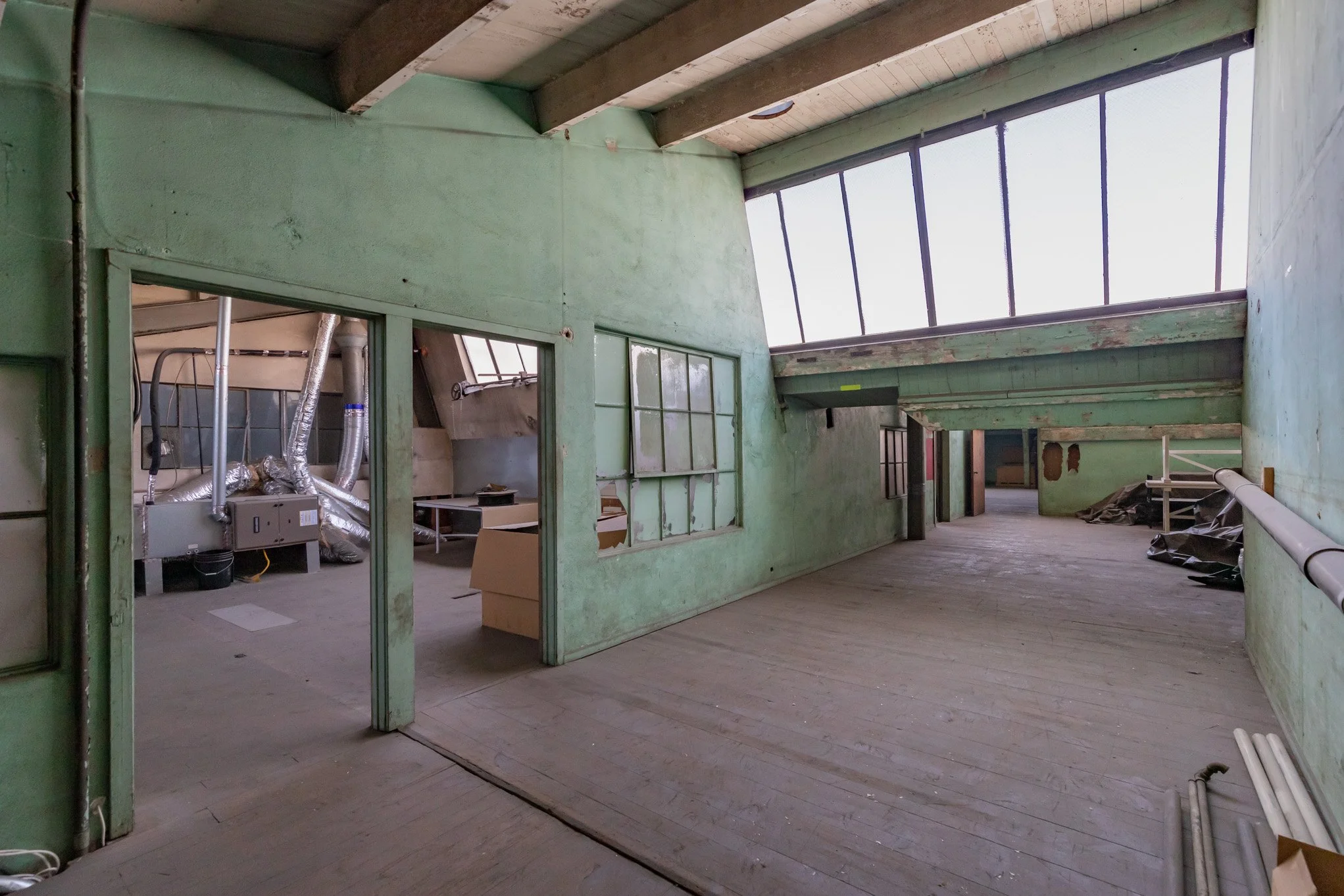 Empty, unfinished indoor space with green walls, large slanted windows, and exposed ceiling beams. Construction materials and black plastic bags are scattered on the floor.