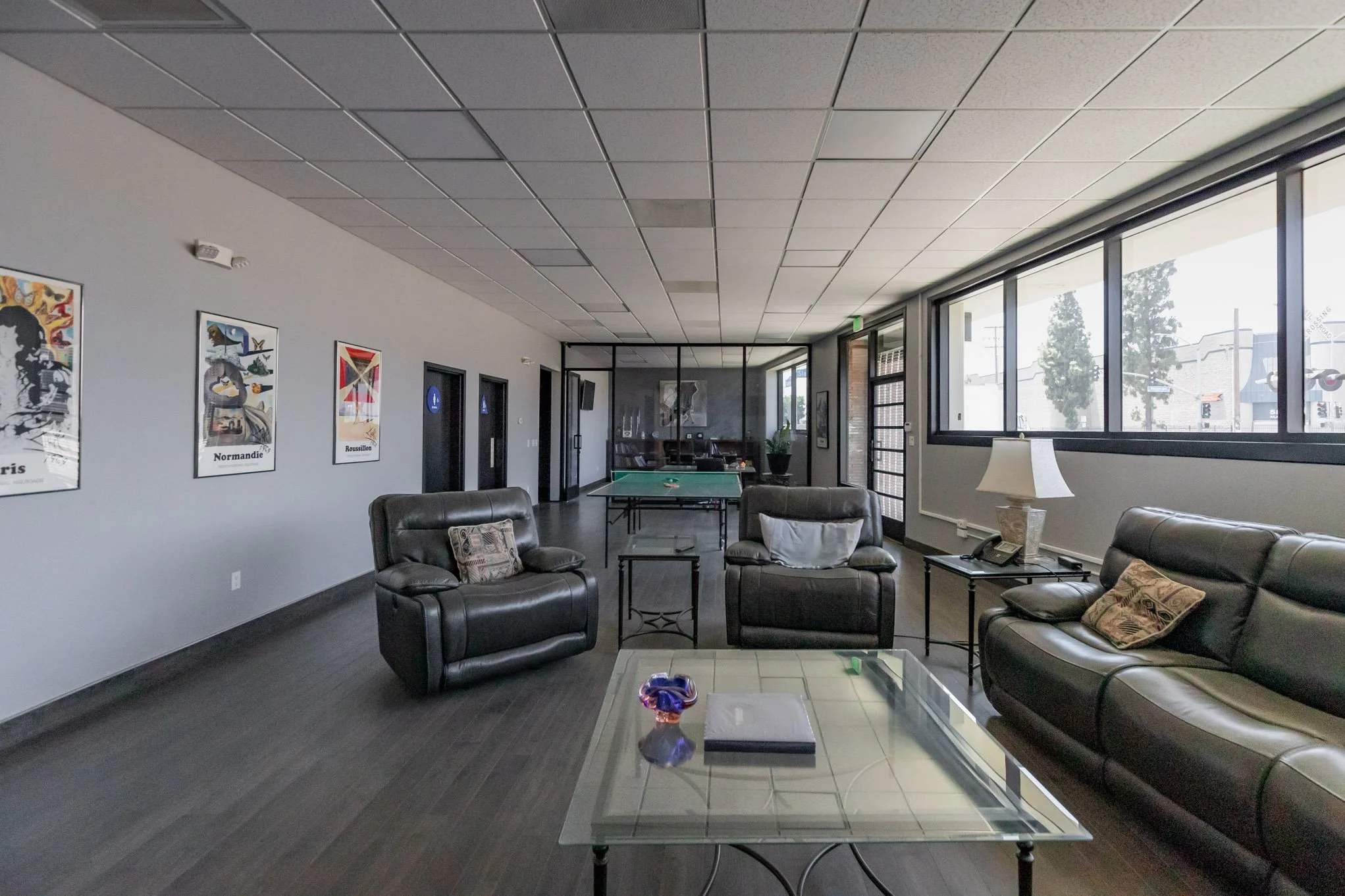 Indoor lounge area with black leather sofas, glass coffee table, and large windows. There is a lamp on a side table, and wall art decorates the wall. A ping pong table is visible in the background.