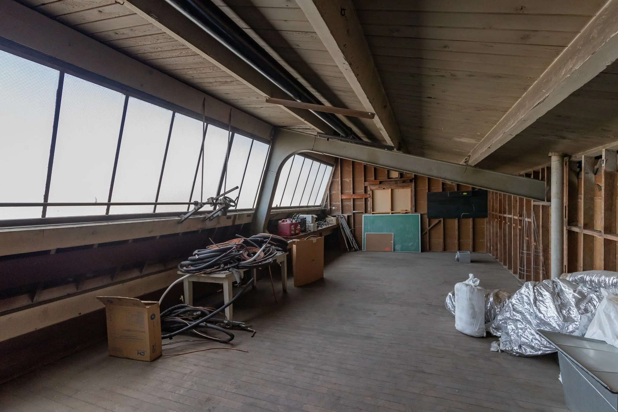 Empty attic with unfinished wood walls and ceiling, large sloped windows, scattered construction materials, bags, and an artificial grass board.