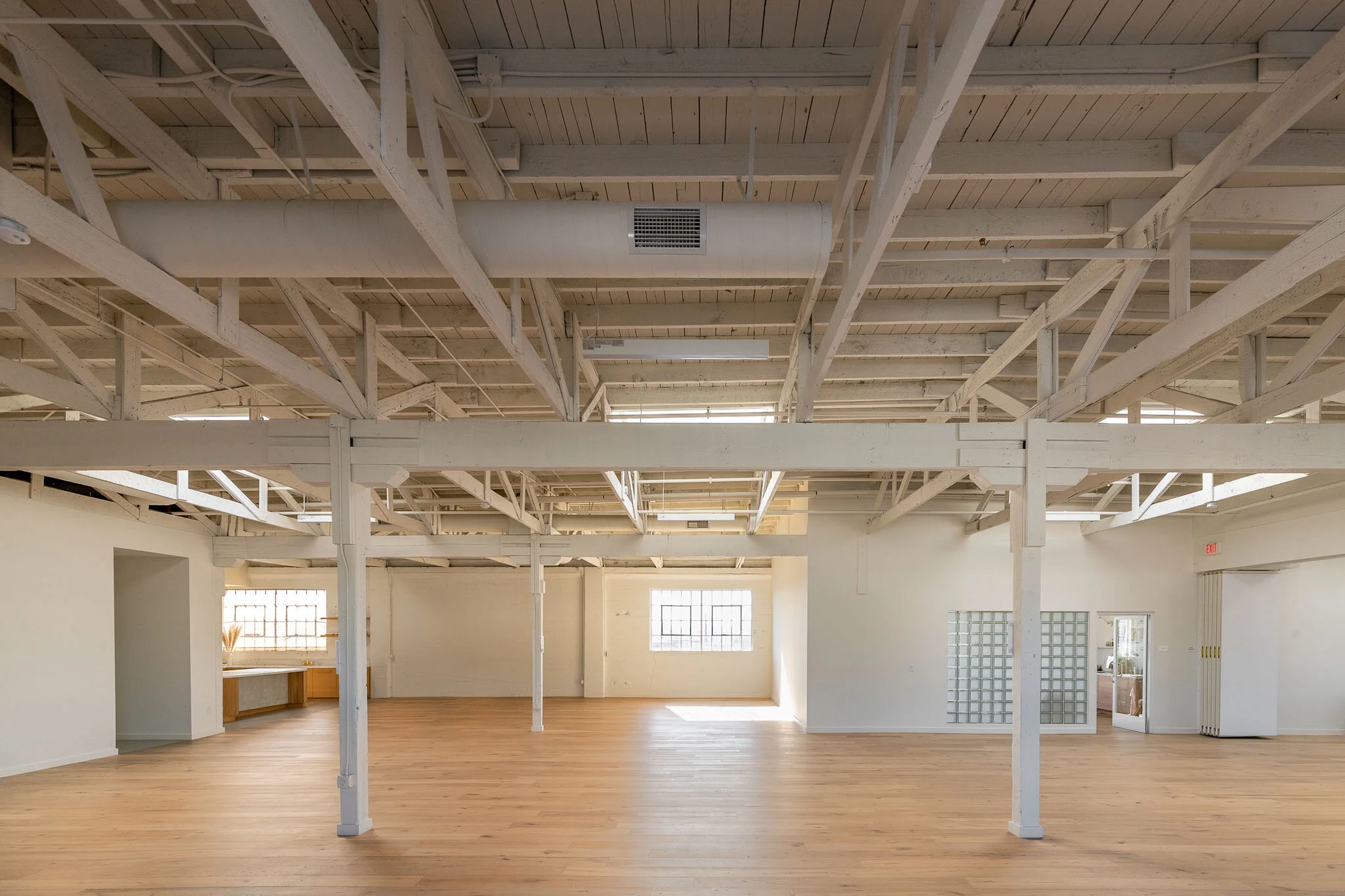 Empty, spacious room with wooden flooring, white walls, large windows, and an exposed ceiling with industrial-style pipes and beams.