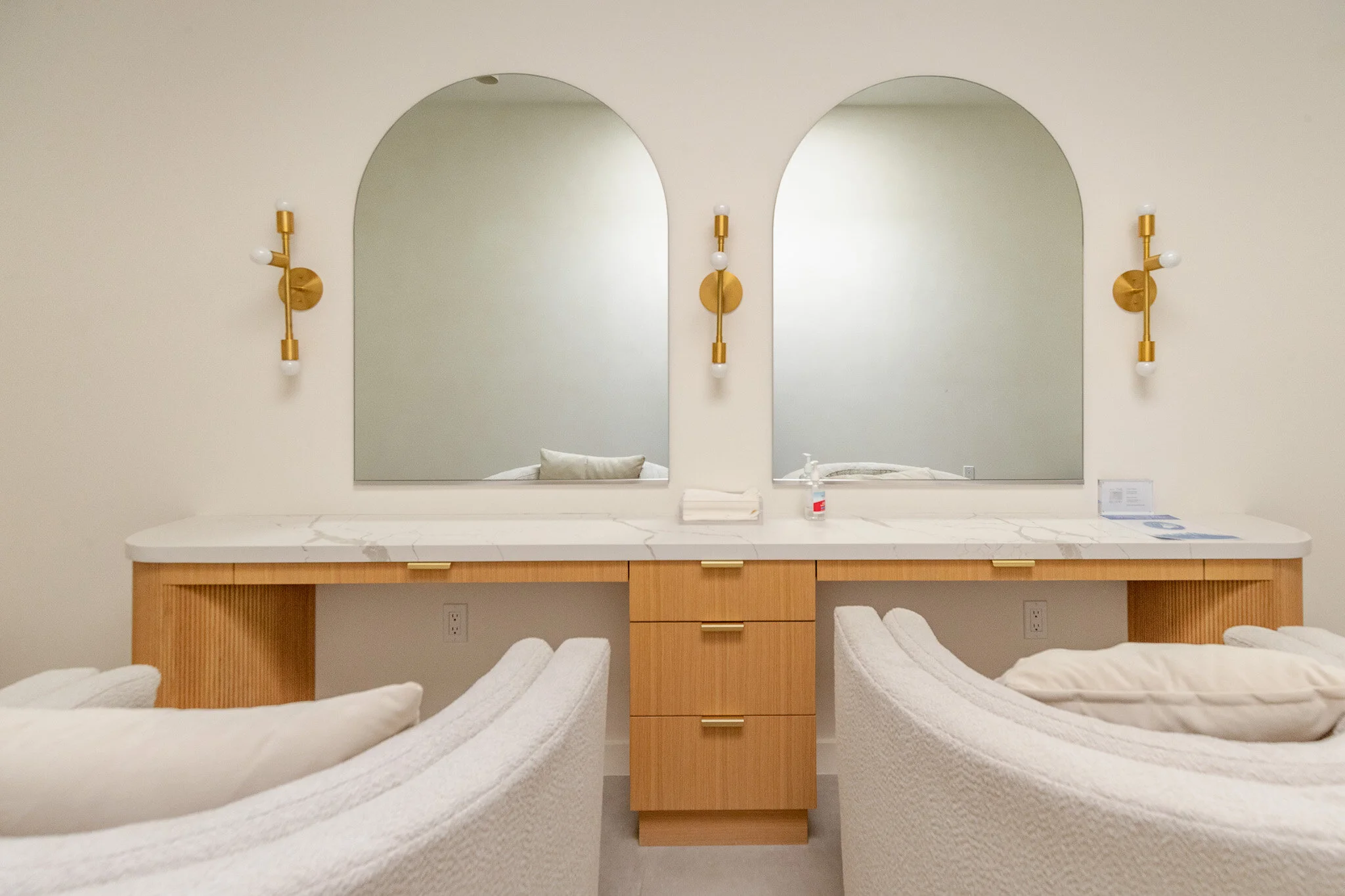 Hotel room vanity with two large mirrors, wooden drawers, white marble countertop, and two modern brass wall sconces. There are white upholstered chairs with pillows in the foreground.