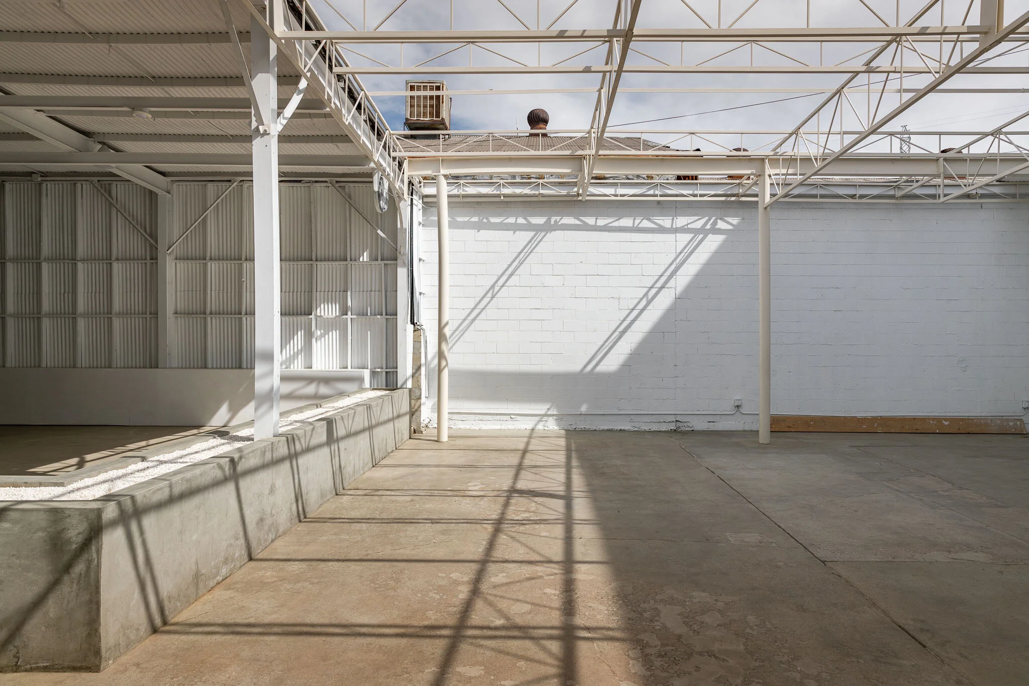Empty outdoor patio area with concrete floor, white brick wall, metal roof structure, and shadows from the roof trusses.