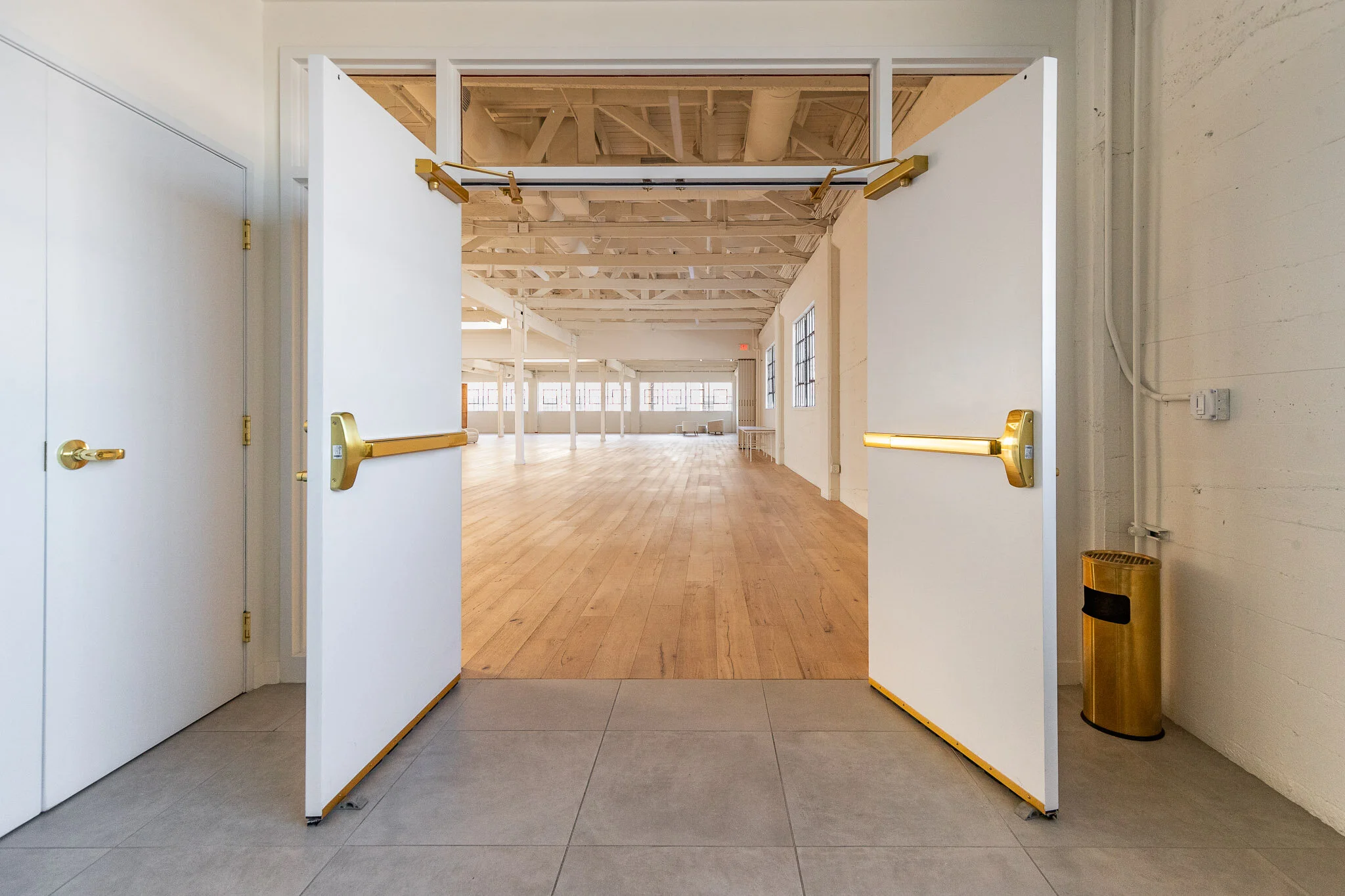 Double glass doors with gold handles open into a large, bright, empty open space with wooden floors and white ceiling beams.