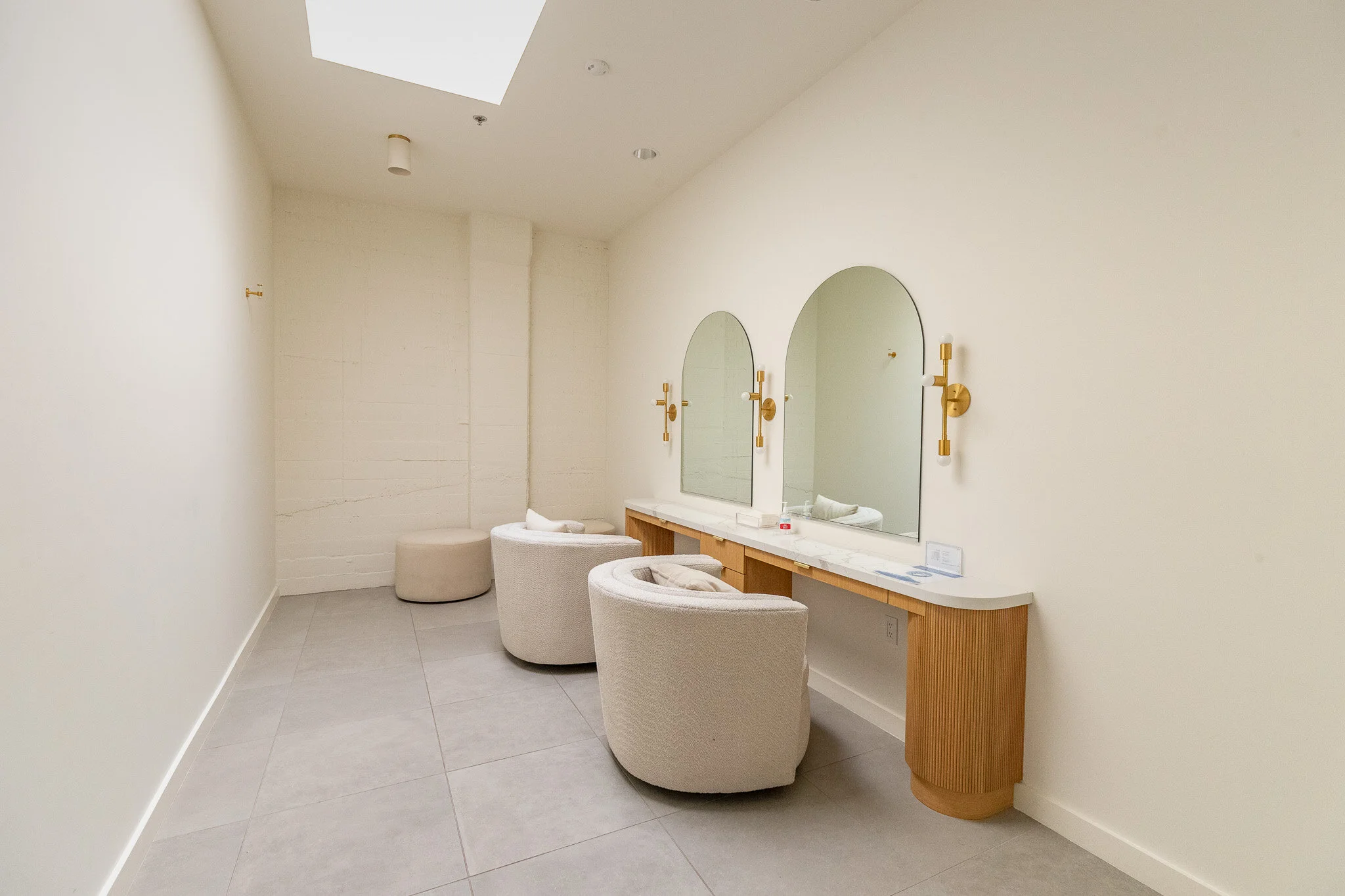 Minimalist bathroom with two arched mirrors, a marble countertop, cream armchairs, and wall-mounted gold light fixtures.