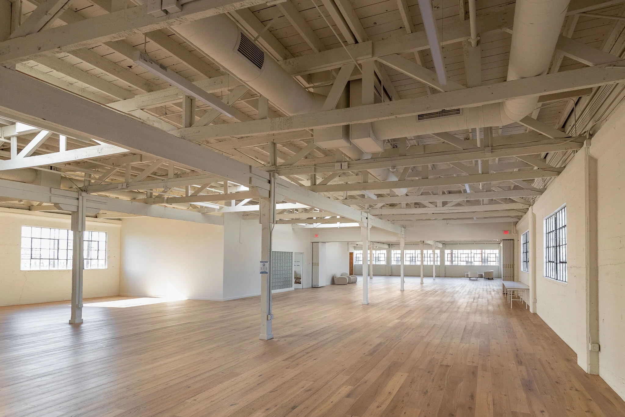 Empty spacious room with wooden floors, white painted ceiling with exposed beams and pipes, large windows on multiple walls, and a few small tables along the right wall.