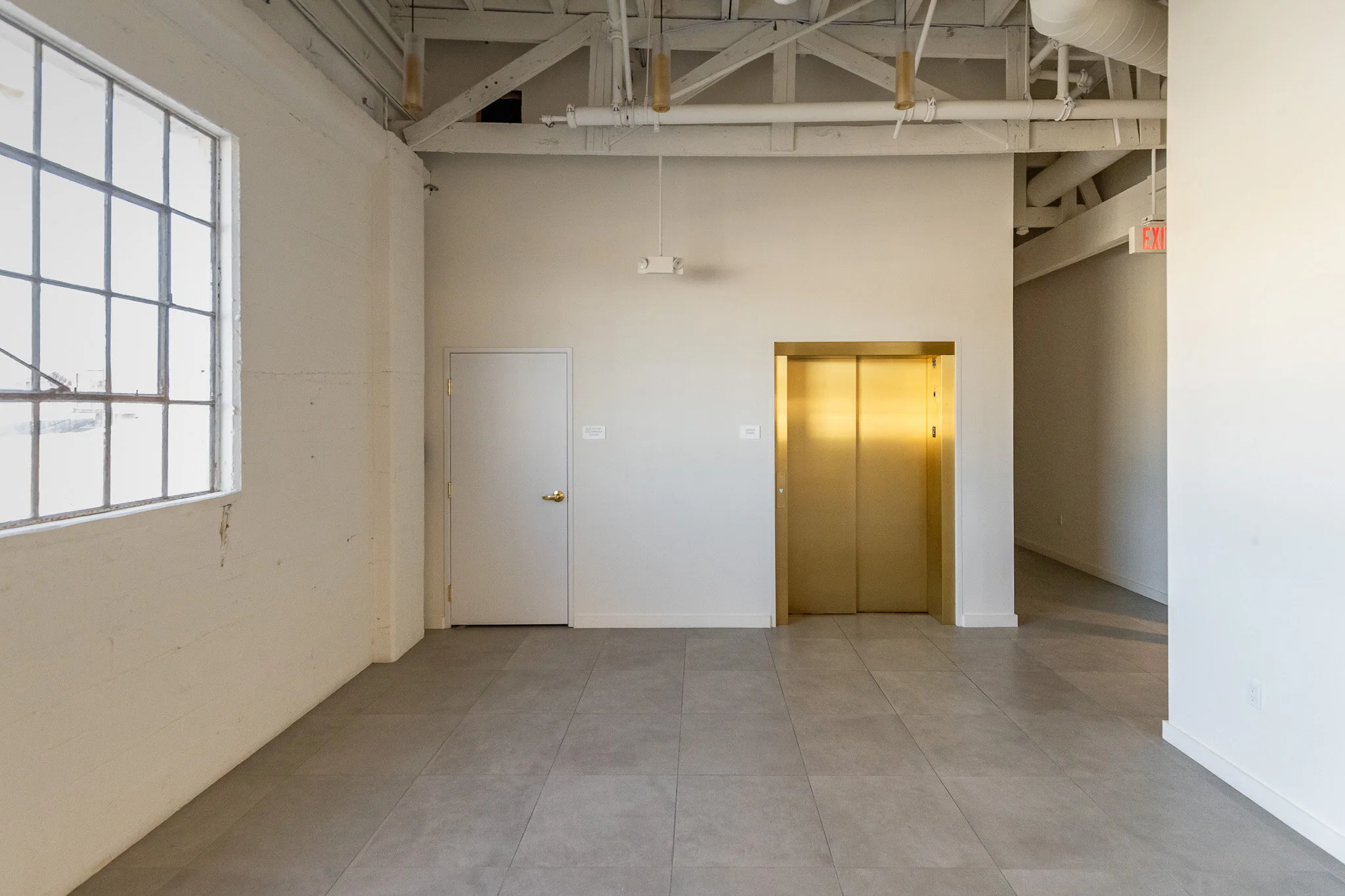 Empty room with large window, white walls, gray tile floor, gold elevator doors, white door, visible ceiling pipes, and an exit sign.