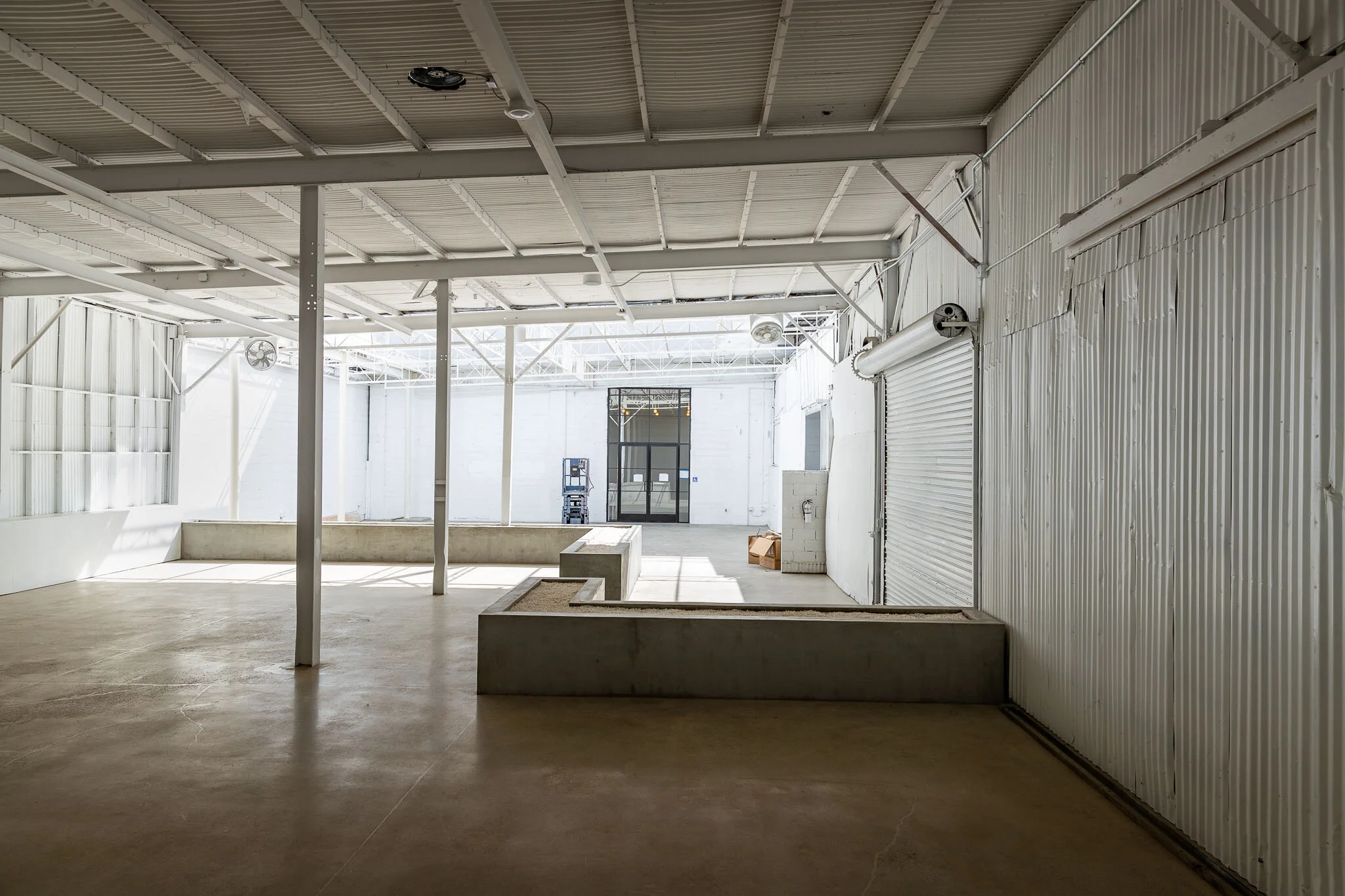 Empty industrial warehouse with concrete floor, metal walls, and ceiling, with a large open space and some construction materials near the back.