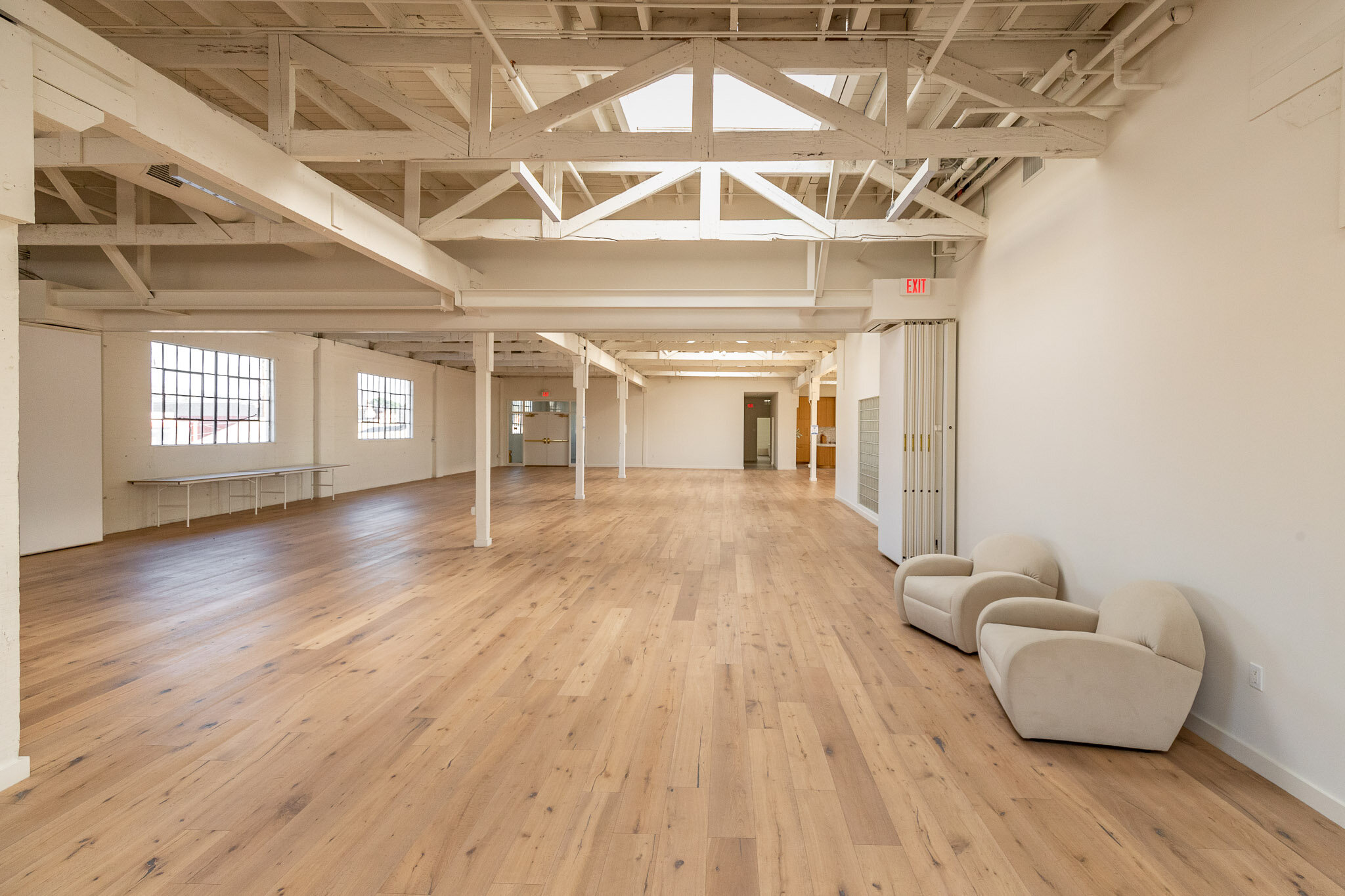 Empty spacious room with white painted exposed ceiling beams, hardwood floors, large windows, and a few beige armchairs in the right corner.