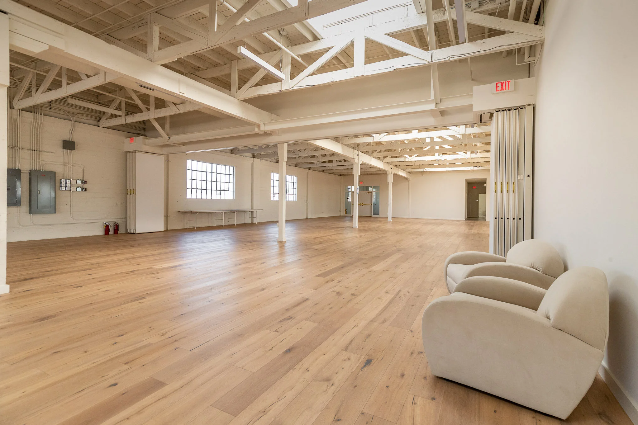 Empty spacious room with wooden flooring, white walls, exposed ceiling beams, large windows, chairs in the corner, and an exit sign.