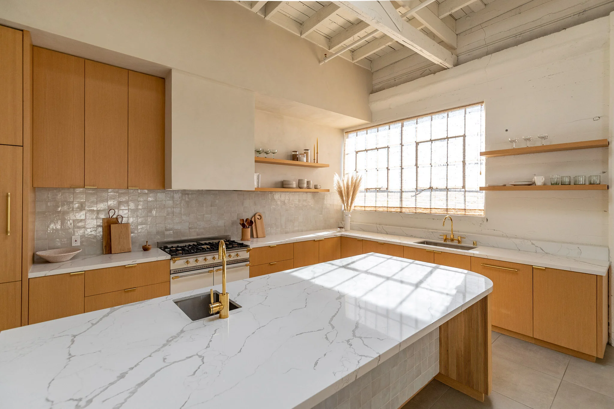 Modern kitchen with white marble countertops, wooden cabinets, and a large window letting in natural light. Gold fixtures are visible on the sink and faucet, with open shelves displaying glassware and dishes.