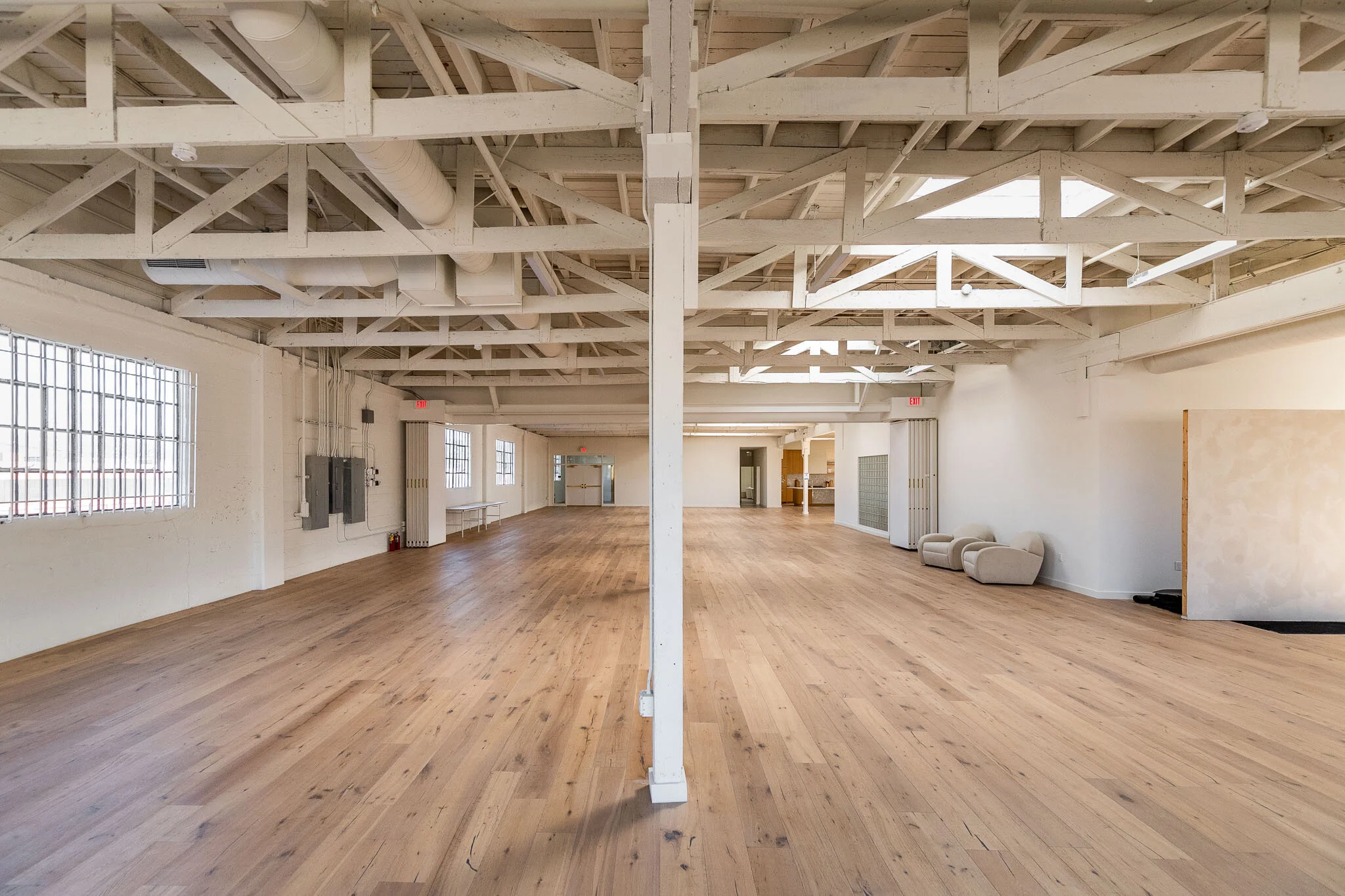 Empty spacious room with exposed white-painted ceiling beams, large windows with grid panes, light wood flooring, and minimal furniture including a few chairs and a table.