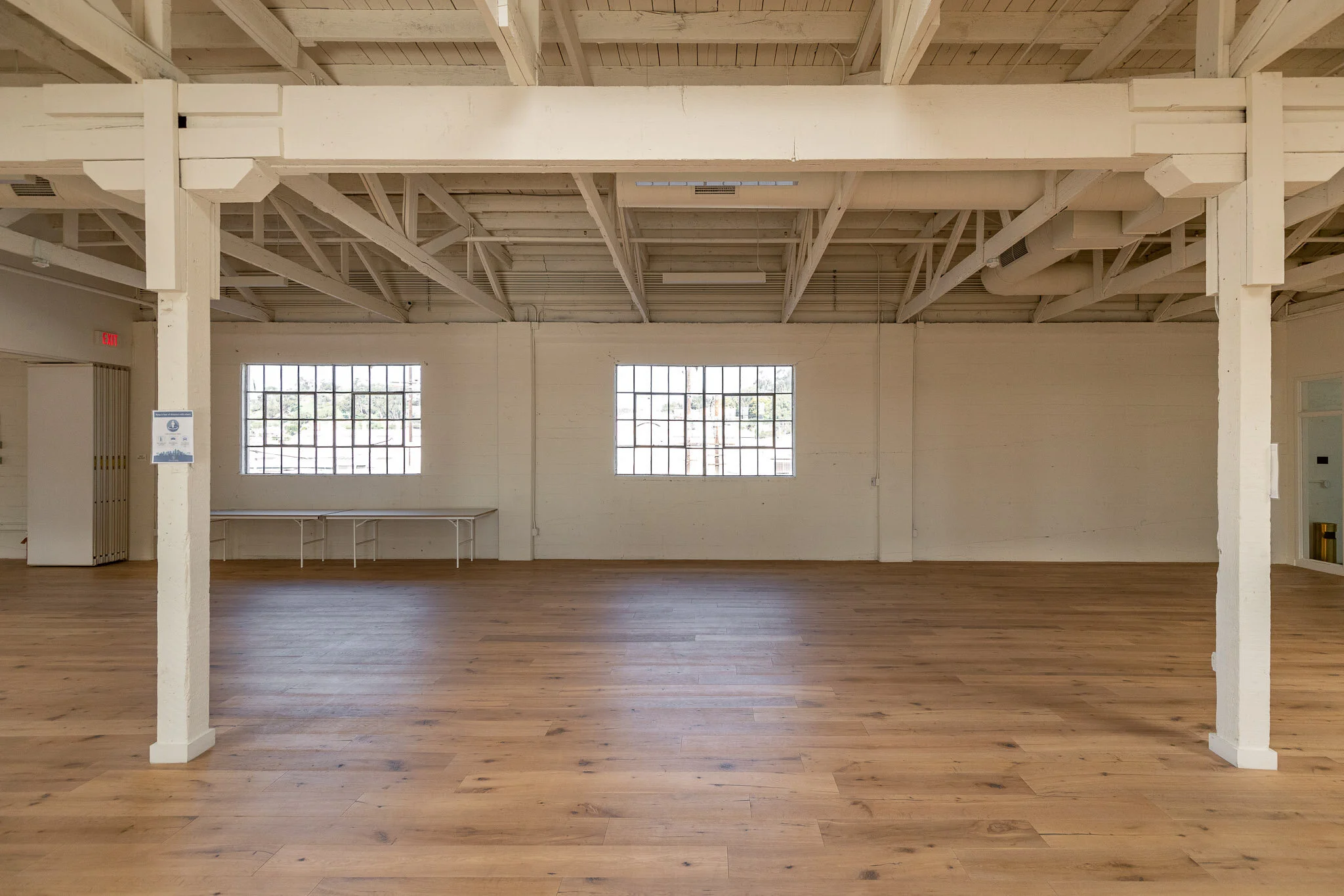 Empty industrial-style room with wooden flooring, white painted brick walls, exposed ceiling beams, two large windows, and a white table along the back wall.
