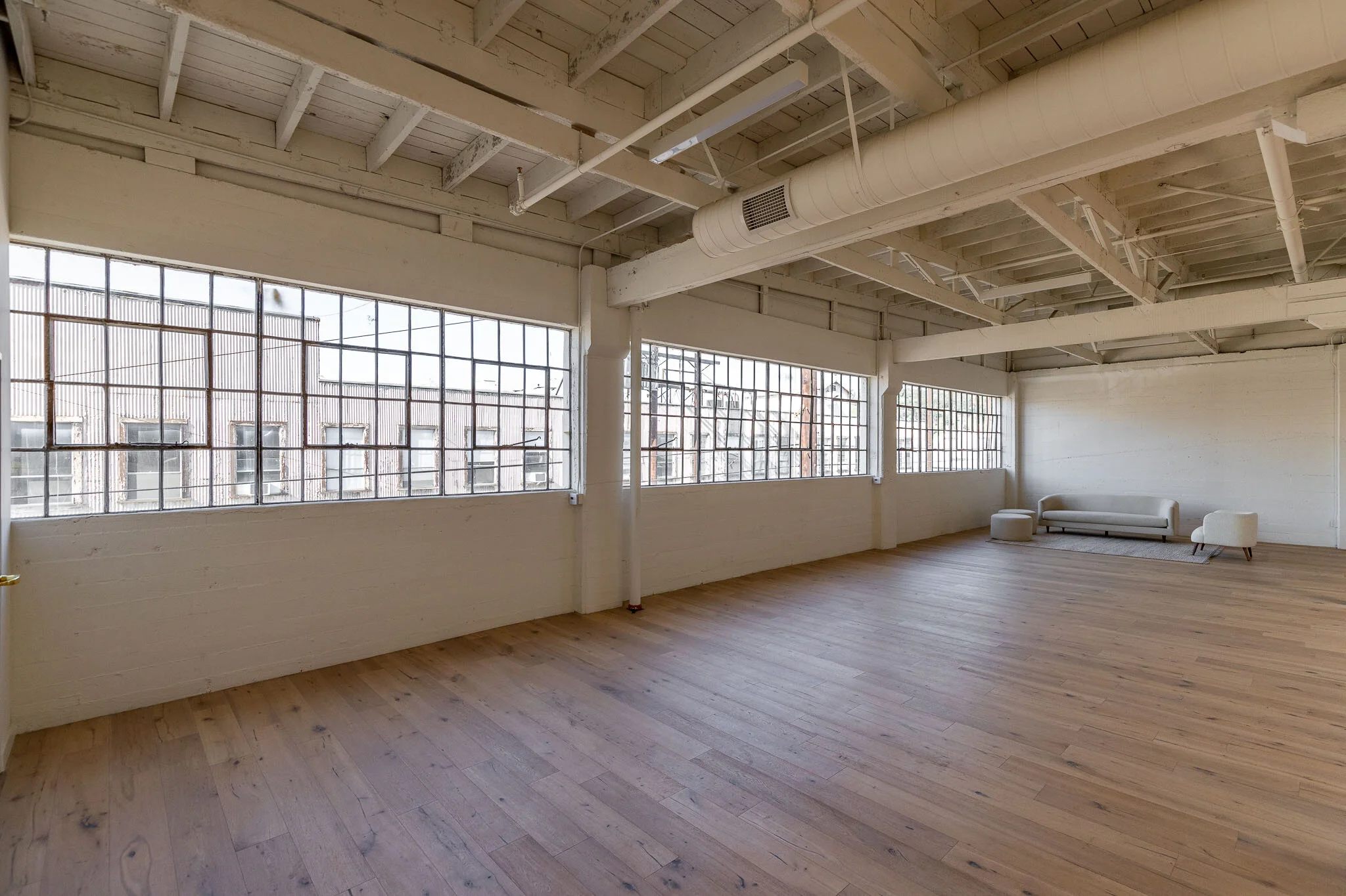 Spacious loft with large industrial-style windows, white walls, exposed ceiling beams, and minimal furniture including a sofa, ottoman, and side table.