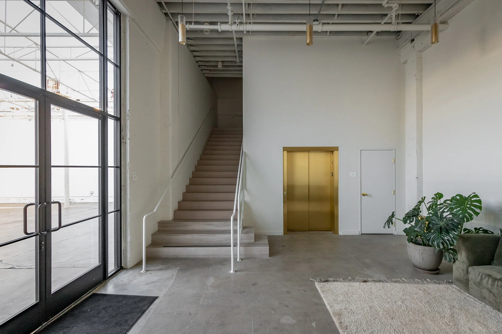 Interior view of a modern minimalistic lobby with a staircase, gold elevator door, white walls, large window, potted plant, and seating area.