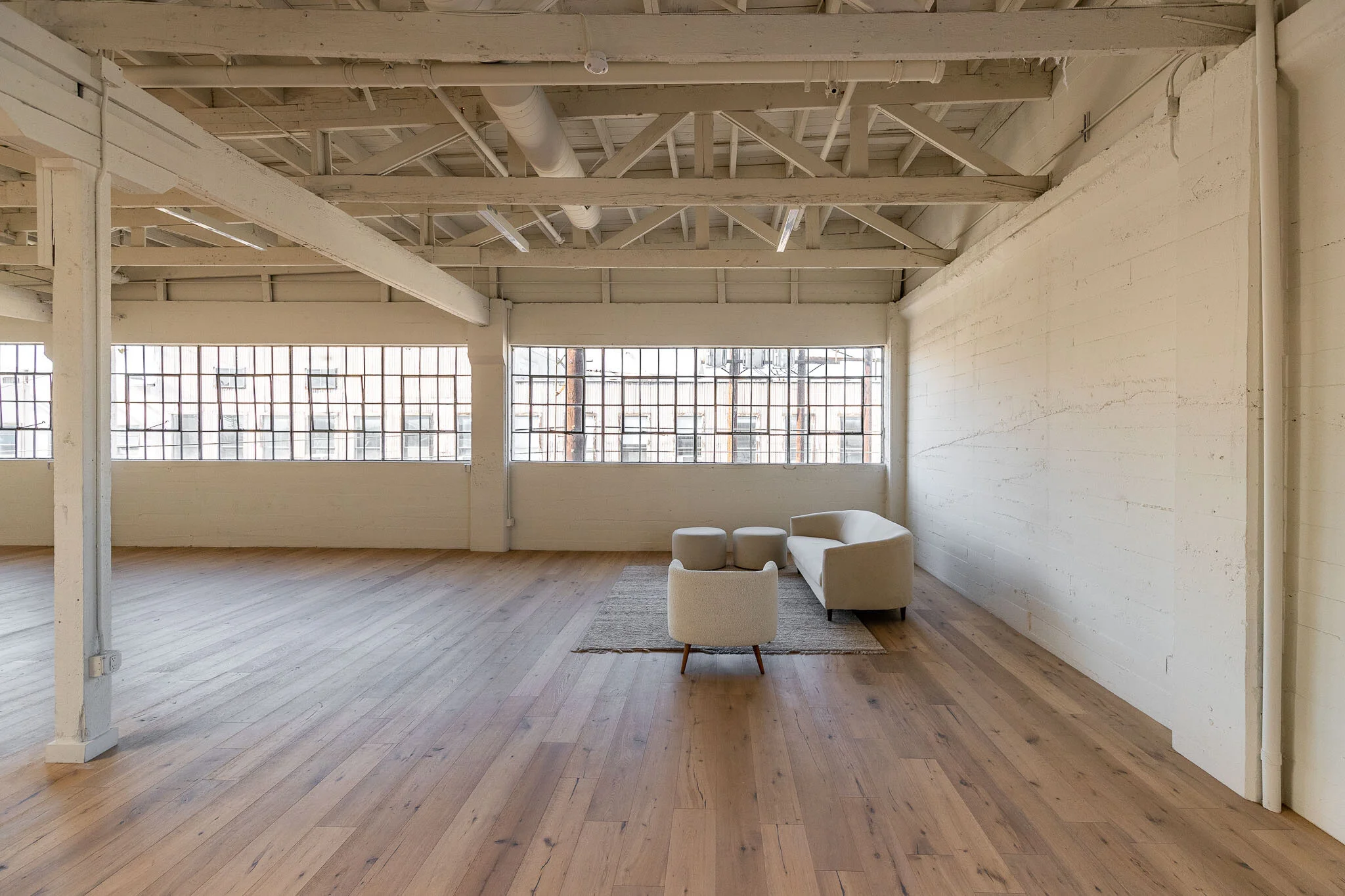 Empty industrial-style loft with wooden flooring, large windows, and minimal furniture including a sofa, armchair, and two stools on a rug.