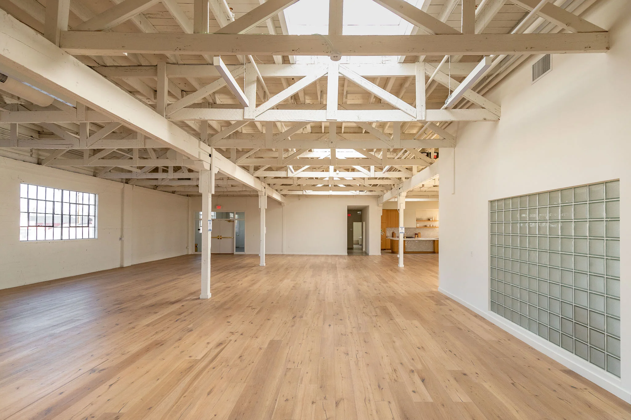 Empty industrial-style open space with wooden flooring, white painted exposed rafters, windows, and kitchen area in the background.