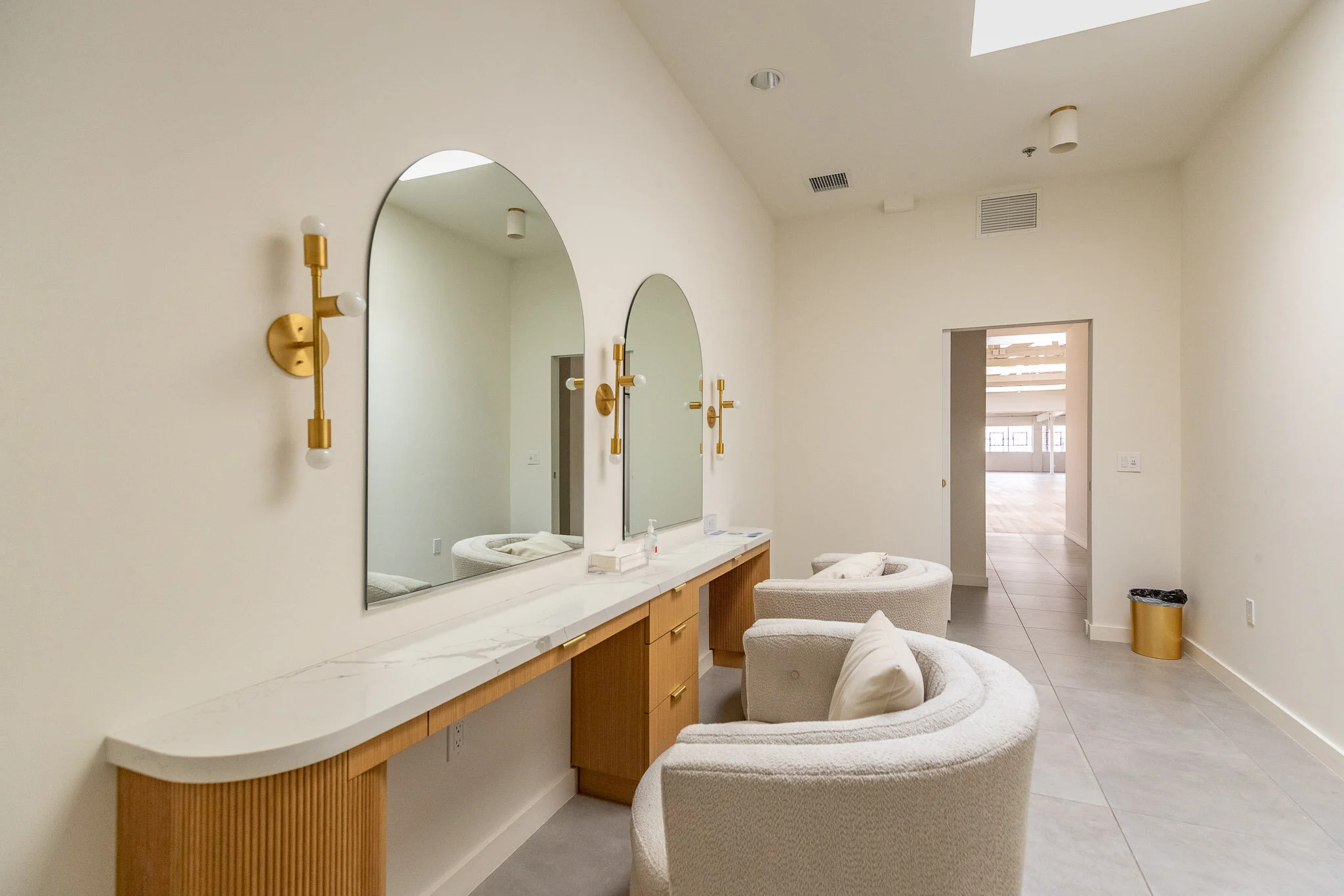 Hair and makeup station with a marble countertop, two large arched mirrors, two white cushioned chairs, gold wall sconces with white bulbs, beige walls, and a doorway leading to a bright room.