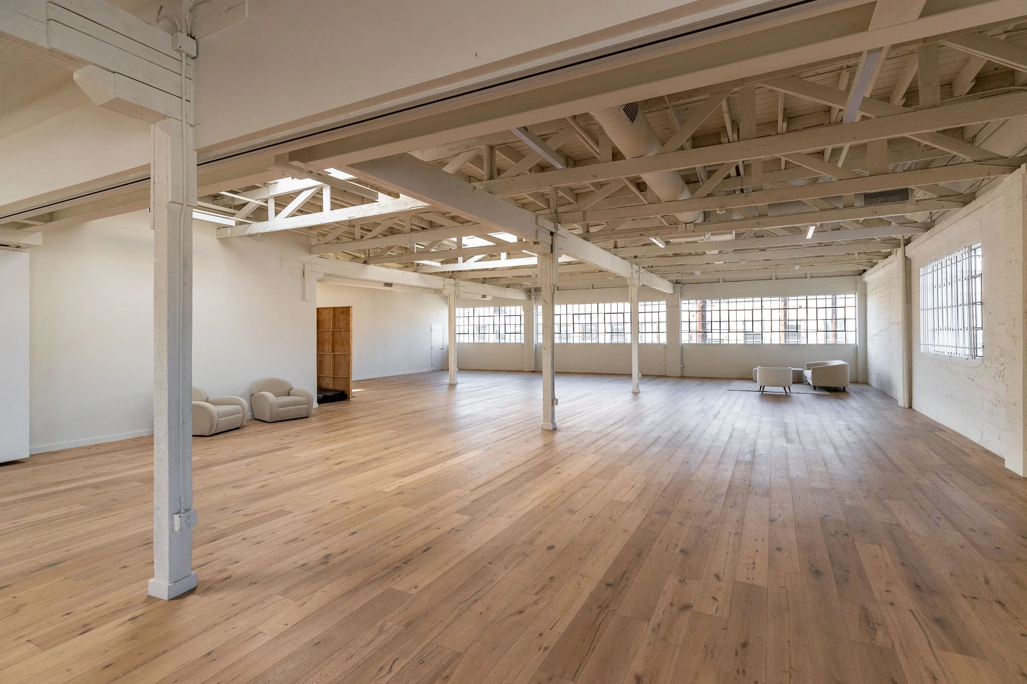 Empty loft with wooden floors, white painted walls, exposed ceiling beams, large windows, and small white armchairs.
