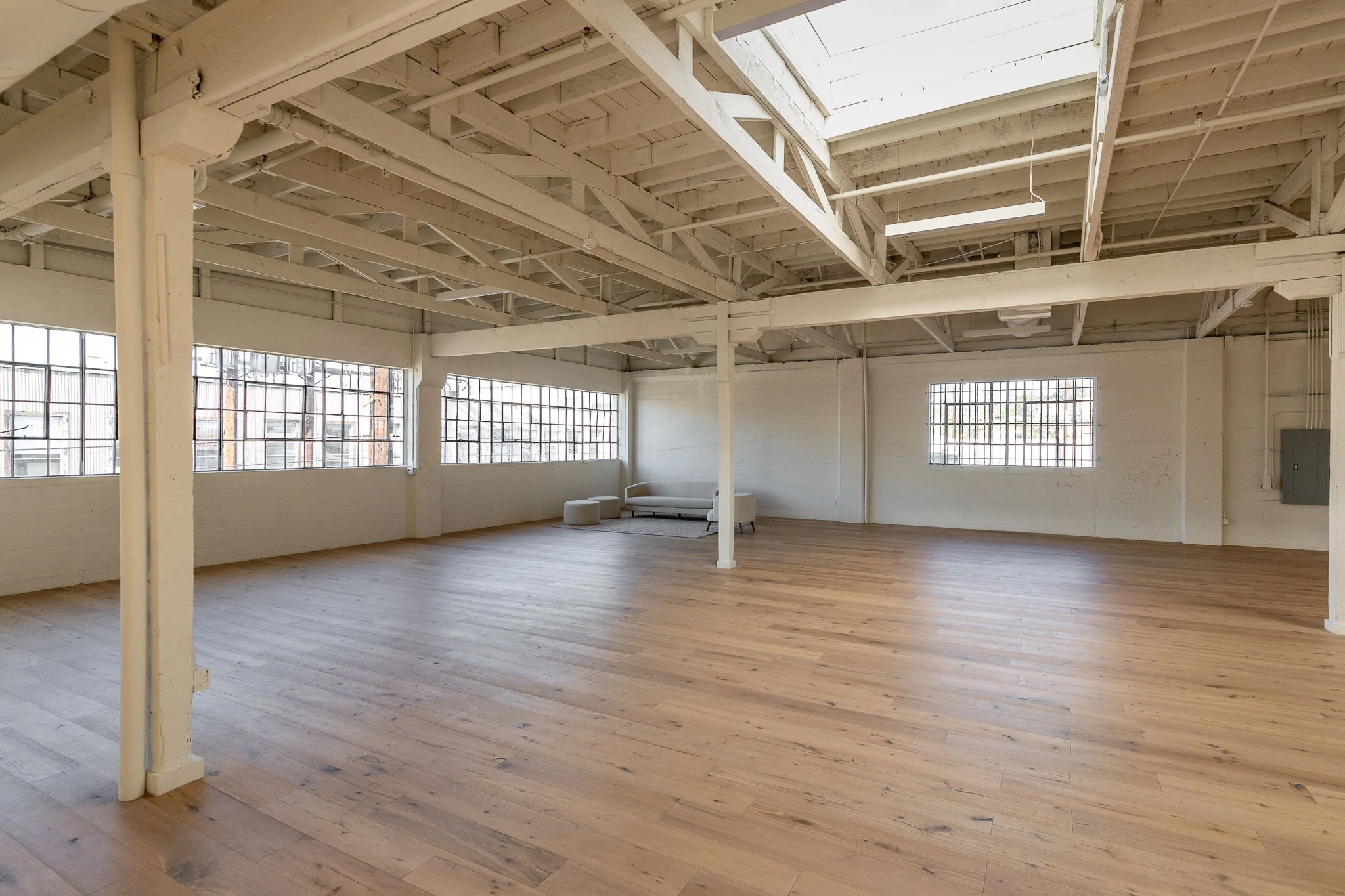 Empty industrial-style room with wooden floors, white painted walls, large windows, and exposed beams and ductwork on the ceiling, with minimal furniture including a sofa and ottomans in the corner.
