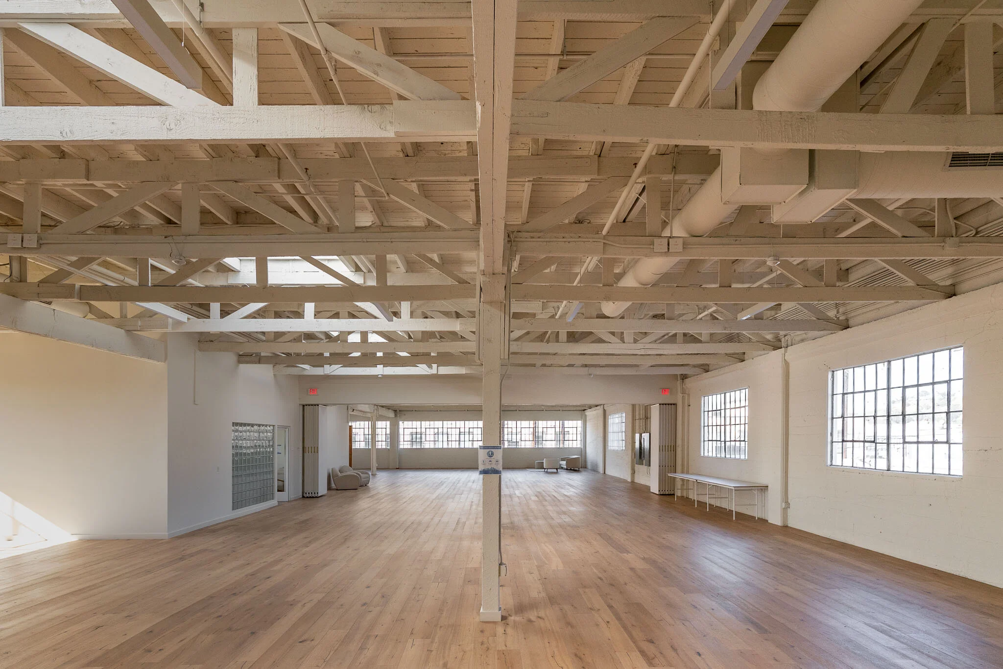Spacious, empty indoor room with large windows, wooden floors, white walls, and exposed ceiling beams.
