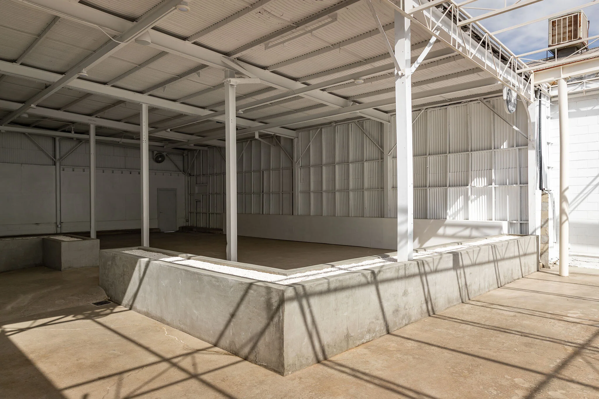 Empty indoor space with concrete floor, white metal walls, and ceiling; structural support beams; shadows cast on the floor from outside light; an industrial environment.