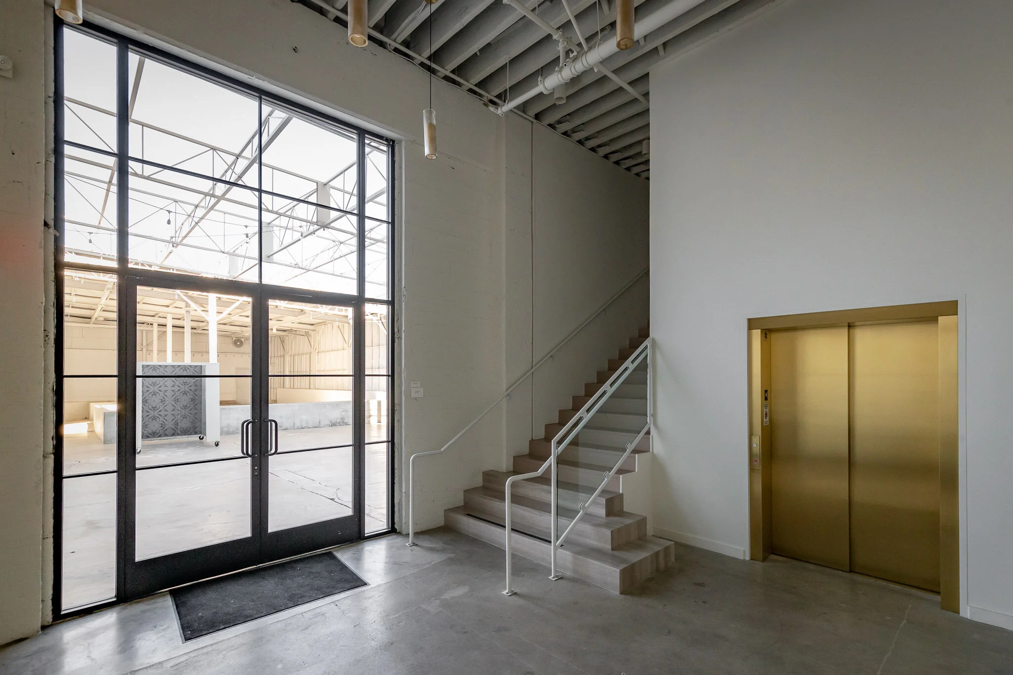 Modern interior with glass door leading outside, wooden stairs with white railings, and an elevator with gold doors in a minimalist design.