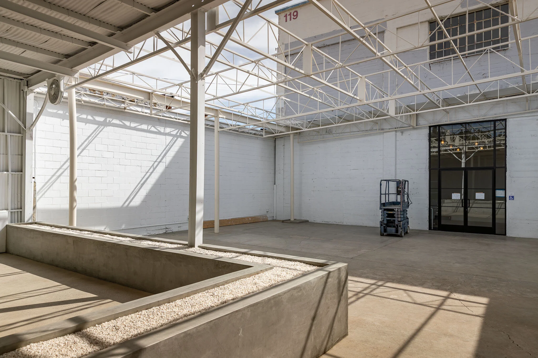 Empty industrial or commercial building interior with white painted brick walls, metal framework, large glass door, and a small lift or scissor jack in the corner.
