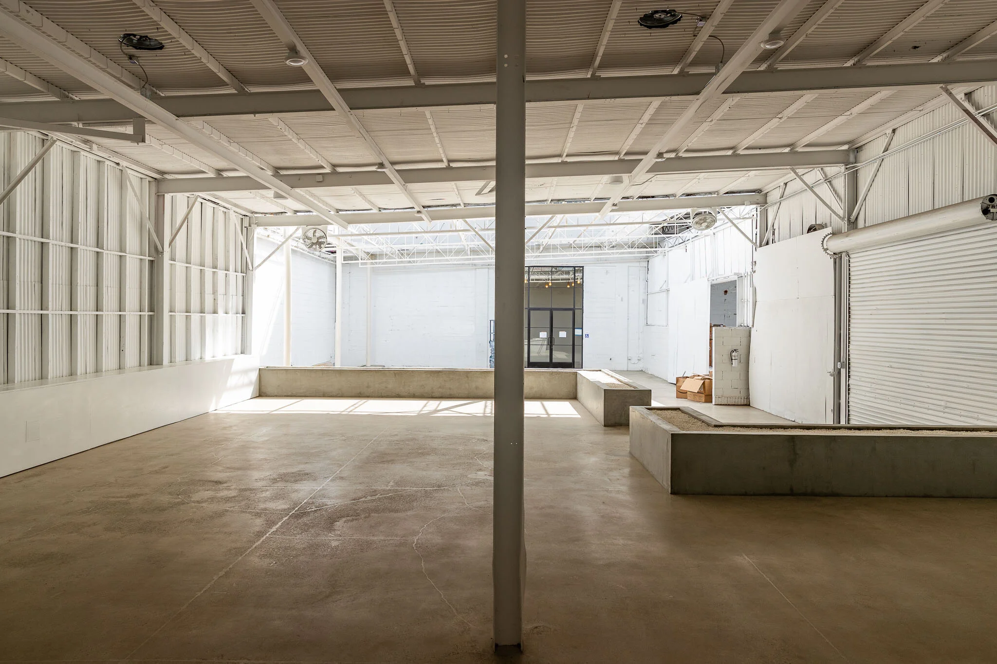 Empty industrial or commercial space with concrete floor, white walls, metal ceiling, and large roll-up door, illuminated by natural light.