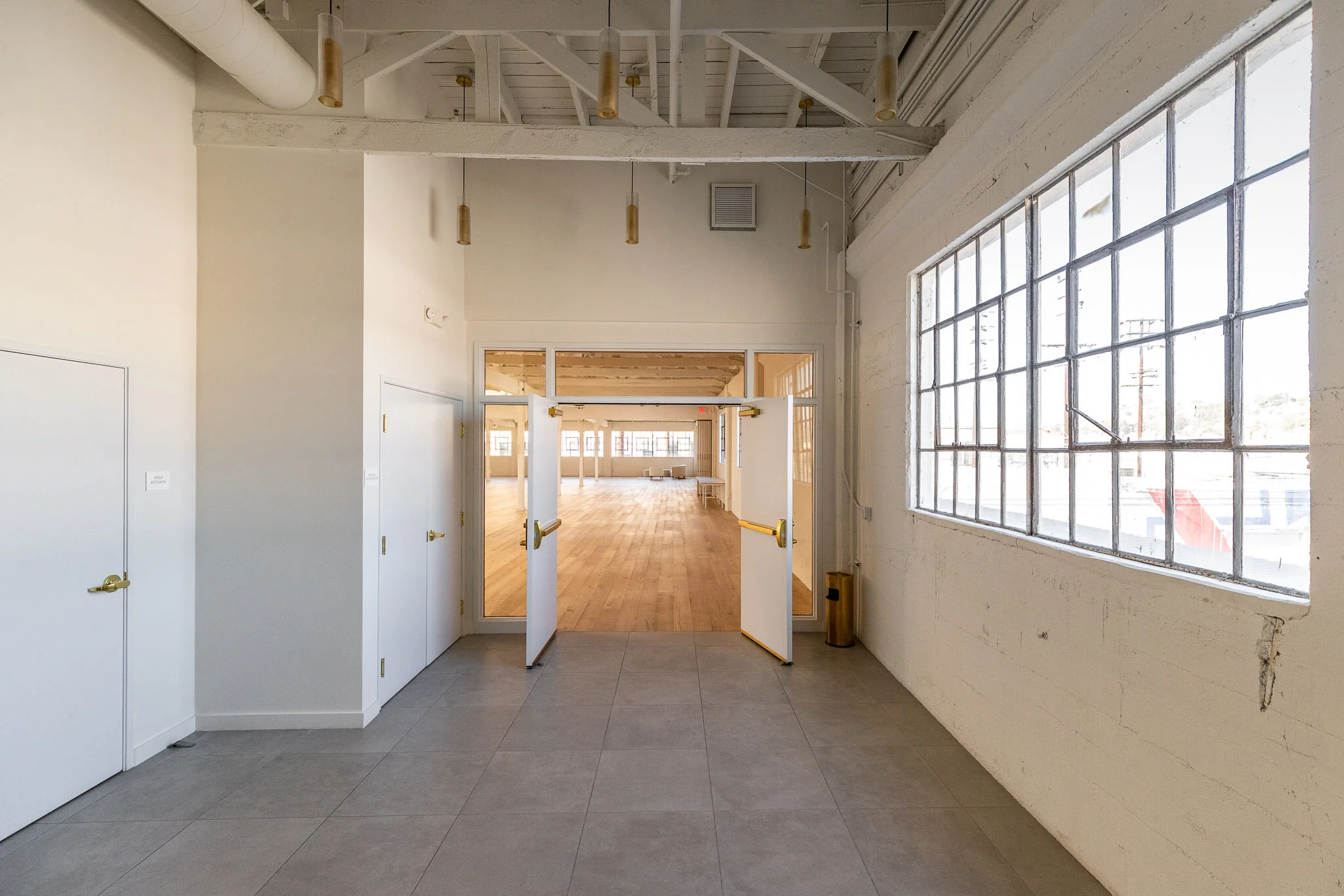 Interior view of an open room with gray tiled floor, white walls, large window on the right, and open double doors leading to a spacious wooden-floored room with support beams and multiple windows.