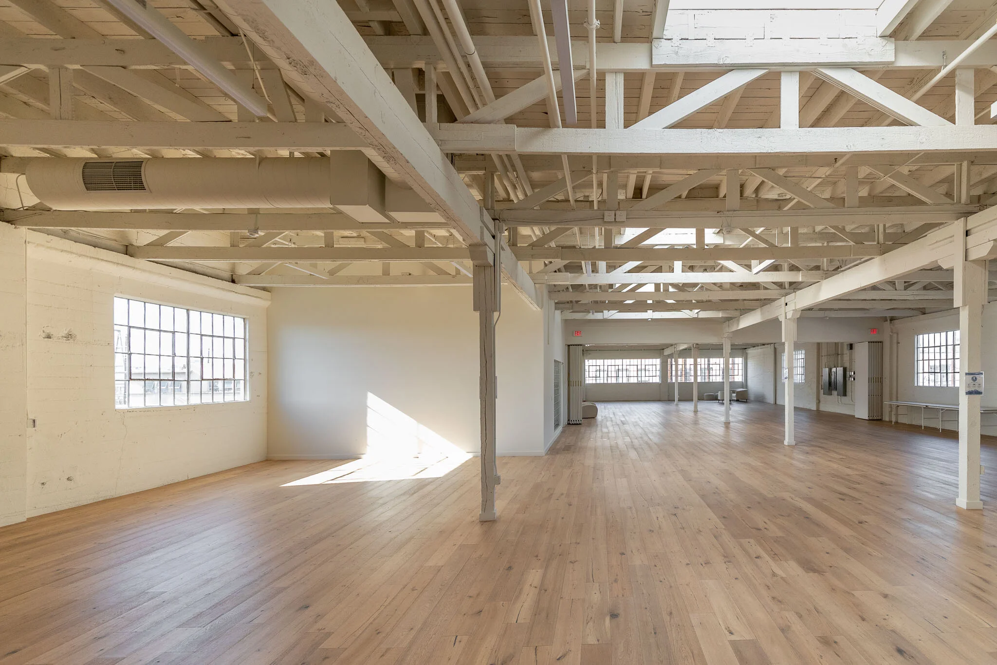 Empty industrial-style interior with large windows, white painted exposed beams, and light wooden flooring.