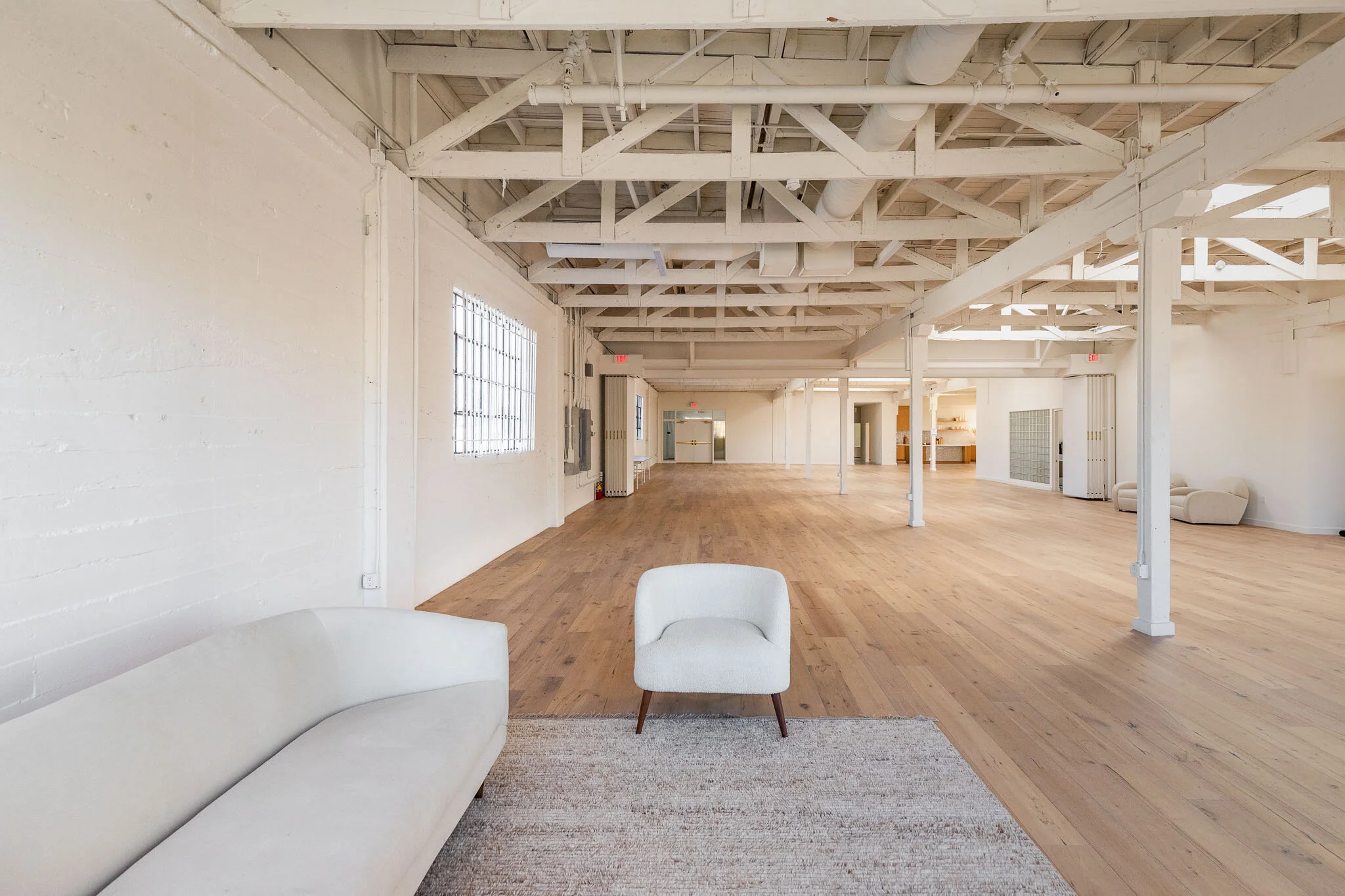 Spacious, minimalist interior with white walls, exposed ceiling beams, wooden flooring, and white furniture, including a sofa and a chair, in an open-plan layout.