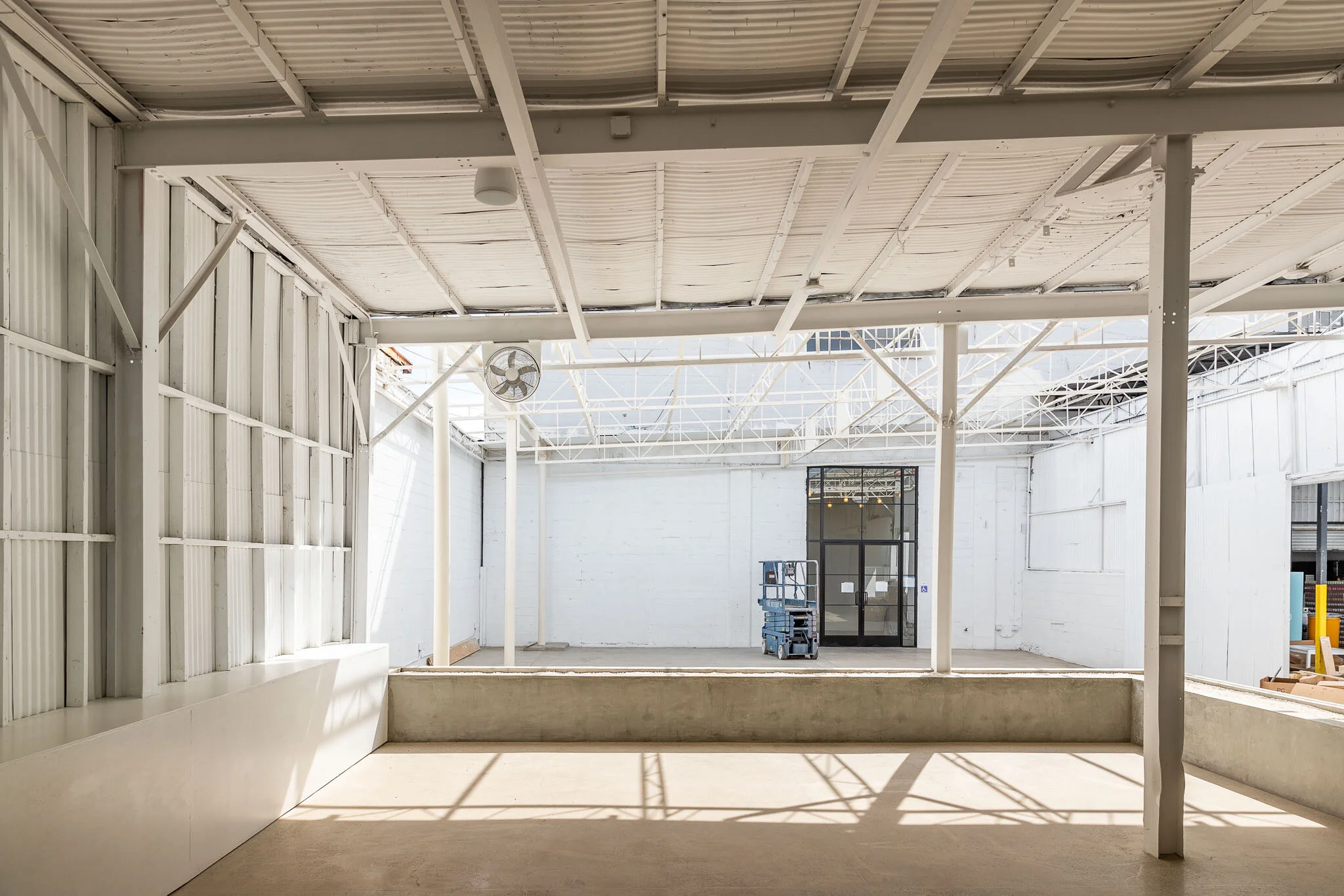 Empty industrial space with white walls, ceiling, and support beams, and a concrete floor. There is a blue lift in front of the back wall and a large window or door with a black frame.