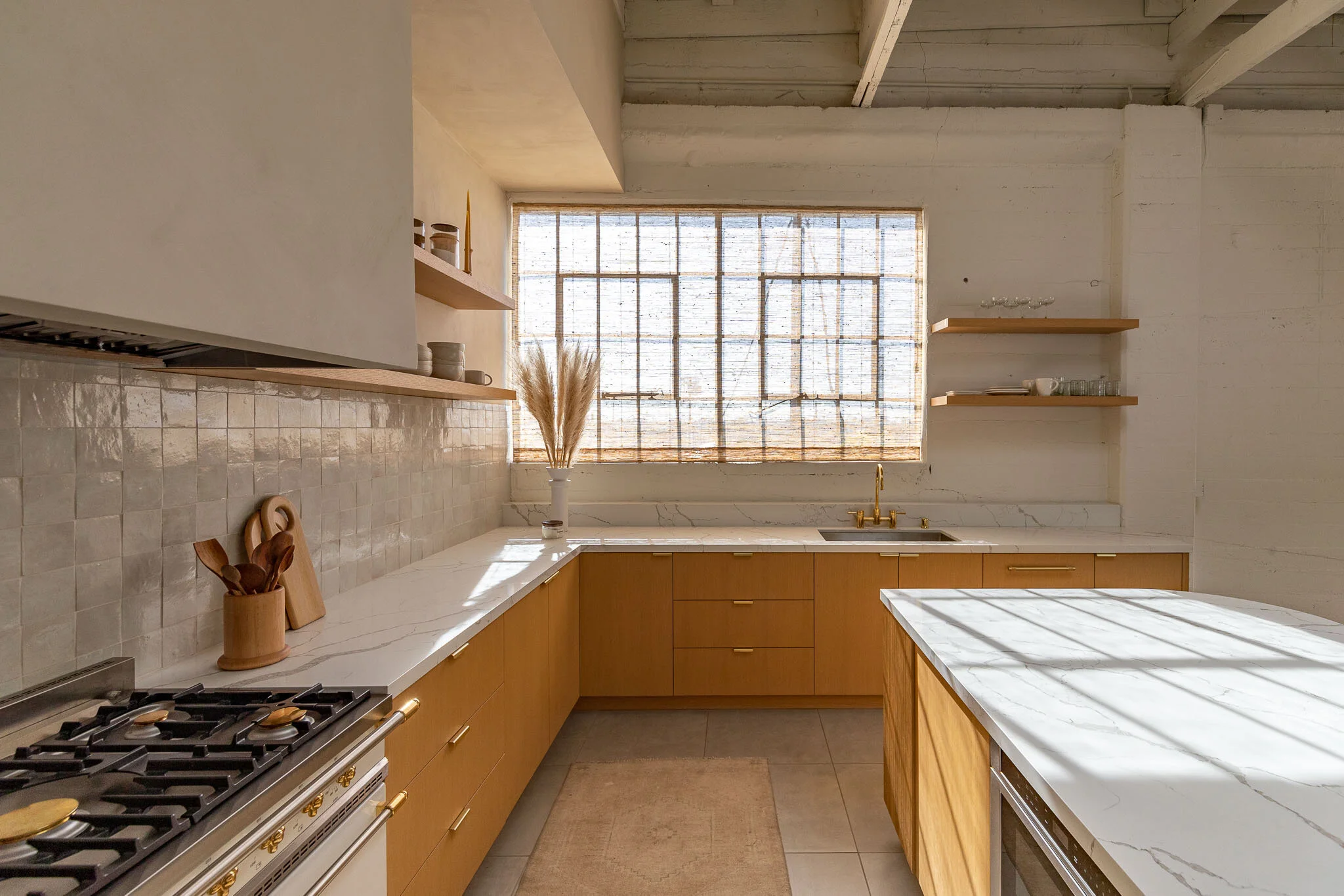 Modern kitchen with white marble countertops, wooden cabinets, and a large window with a textured shade, decorated with a vase of dried pampas grass.