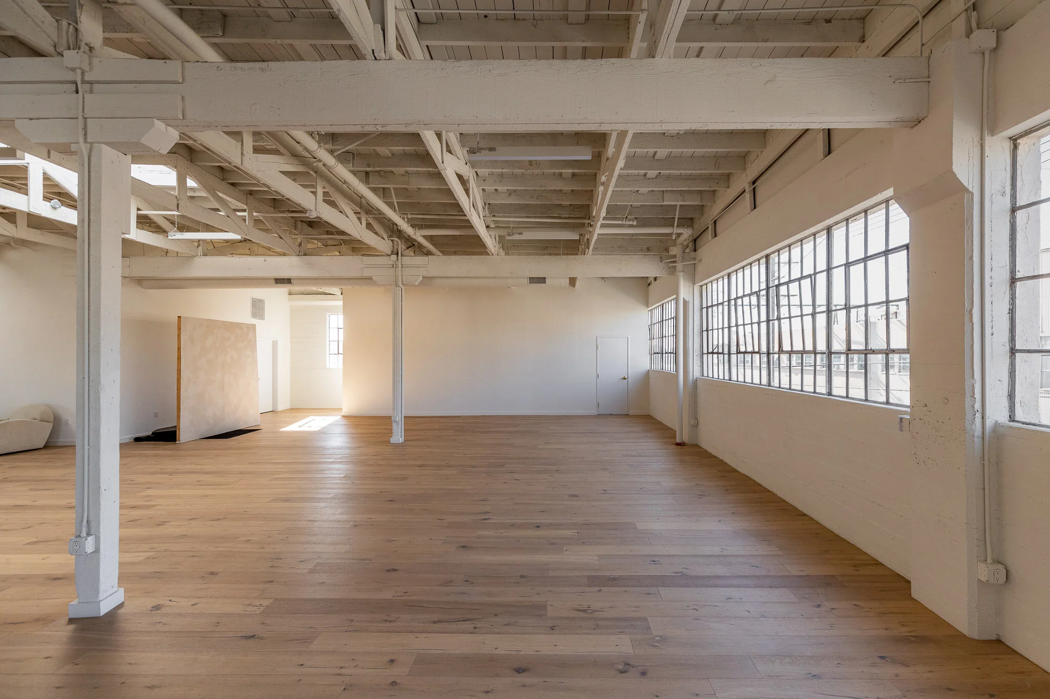 Empty industrial-style room with wooden floor, white painted brick walls, large grid windows, and exposed ceiling beams.