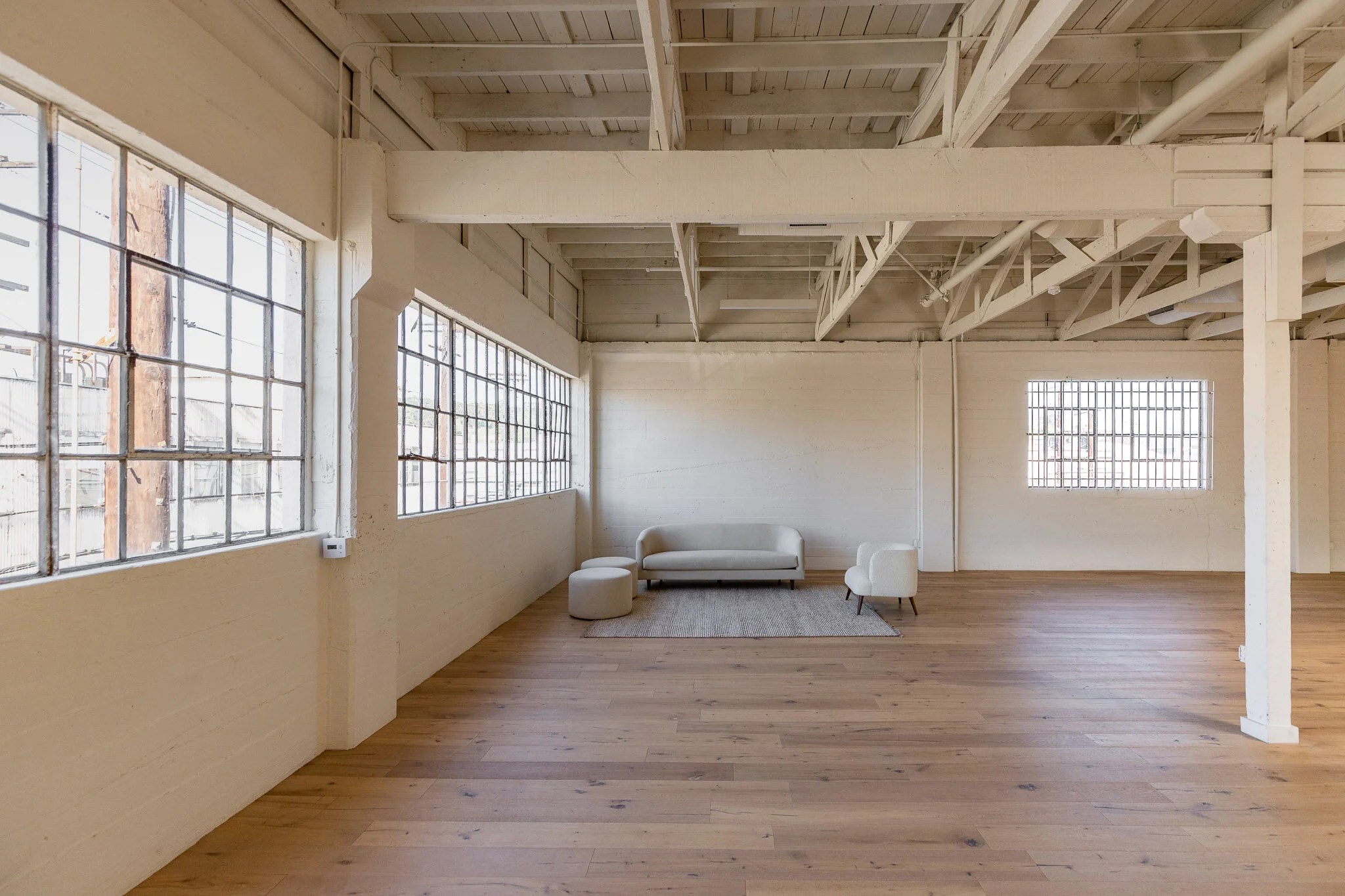 Empty room with white painted brick walls, large industrial-style windows, wooden flooring, and minimalist furniture including a white sofa, a small white armchair, a white ottoman, and a gray rug.