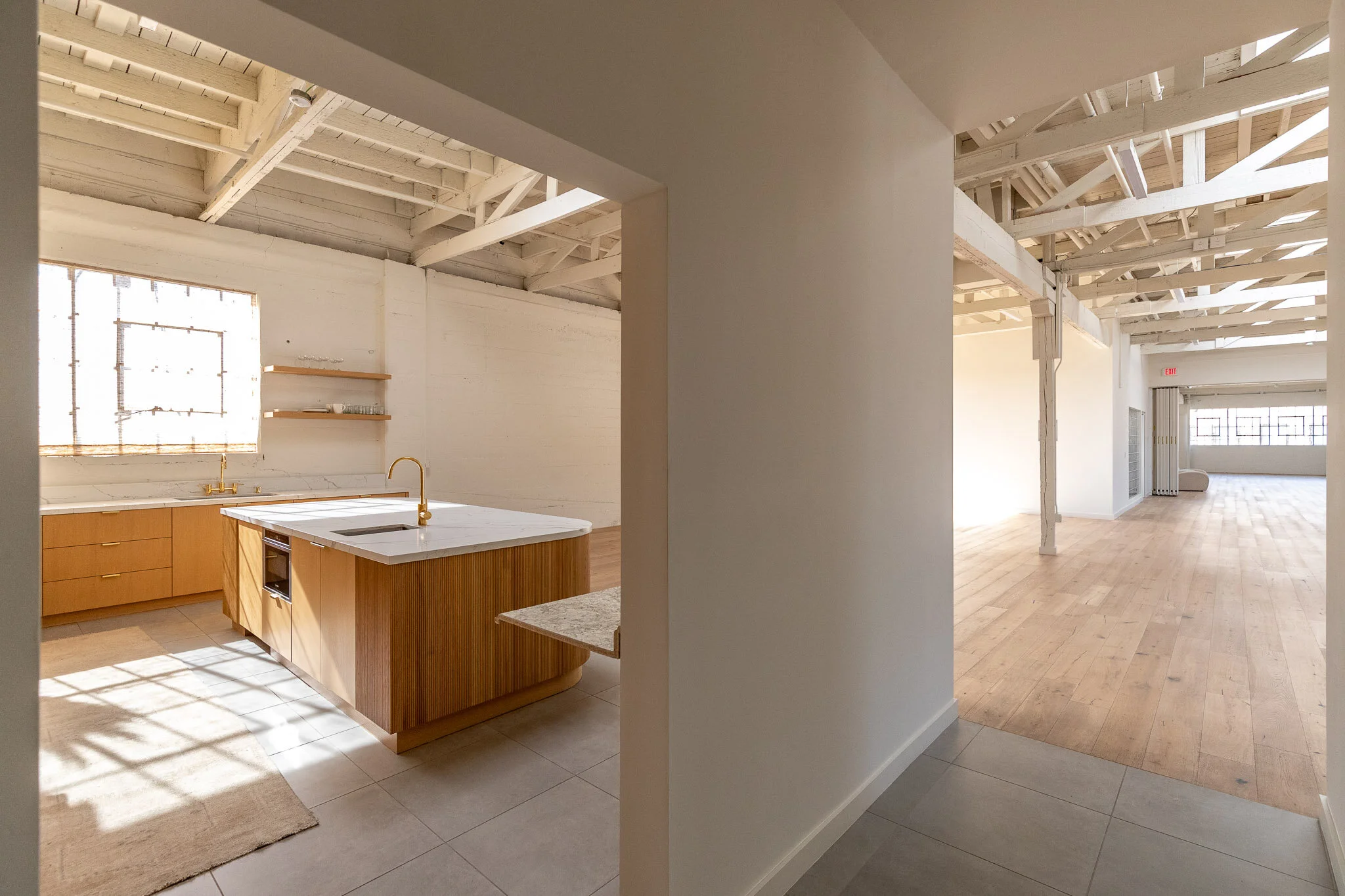 Interior view of a bright, minimally furnished open-plan living space with a kitchen on the left and a large airy room on the right, featuring exposed white ceiling beams and natural light.