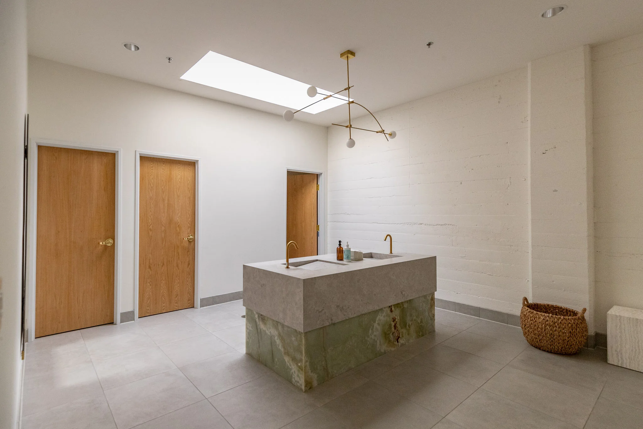 Minimalist bathroom with two wooden doors, a concrete and stone sink with gold faucets, a woven basket in the corner, and a modern chandelier on a white ceiling with a skylight.