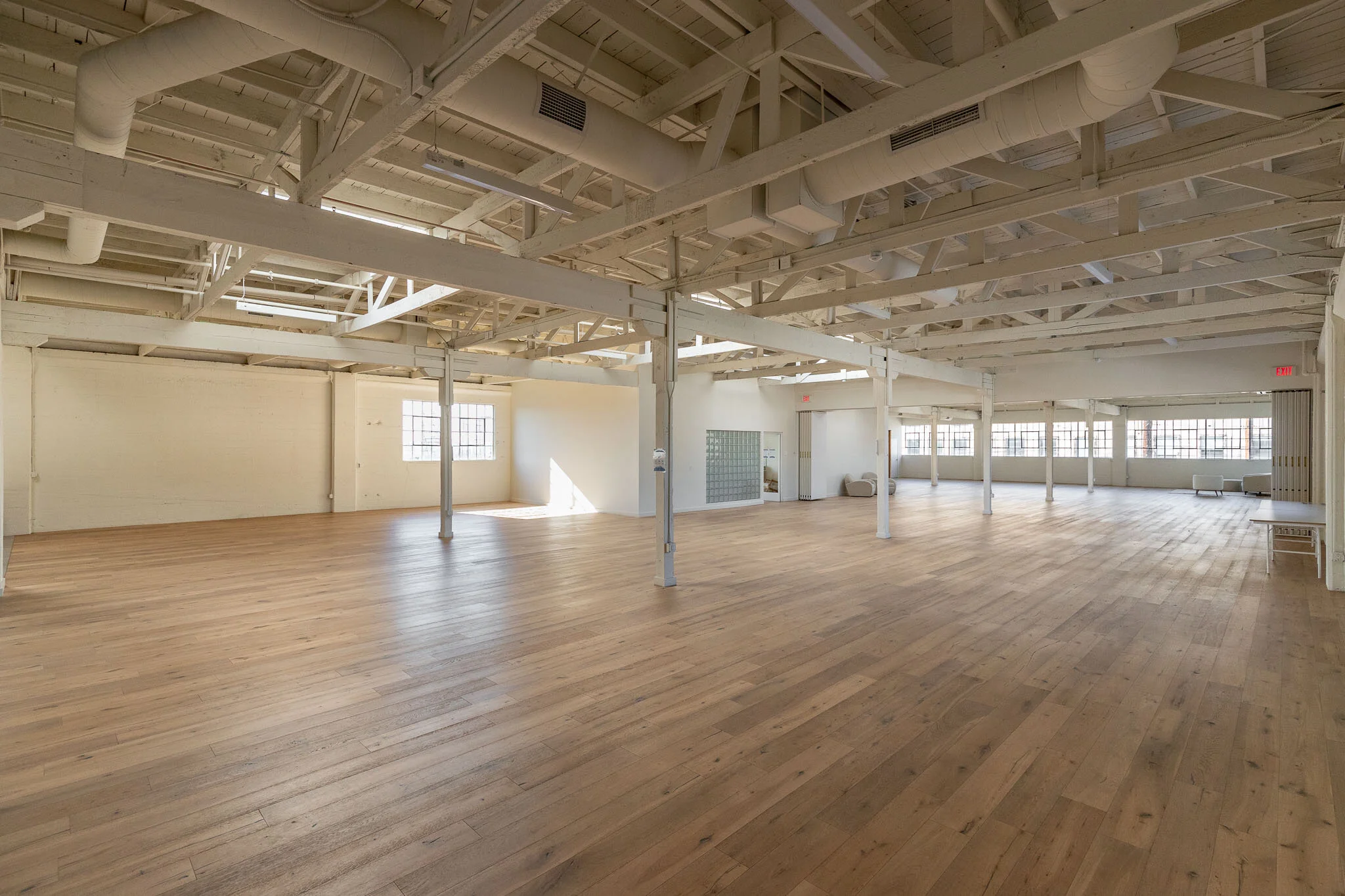 Empty spacious room with wooden flooring, white painted ceiling with exposed beams and ductwork, multiple large windows letting in natural light, and minimal furniture including a few chairs on the right side.