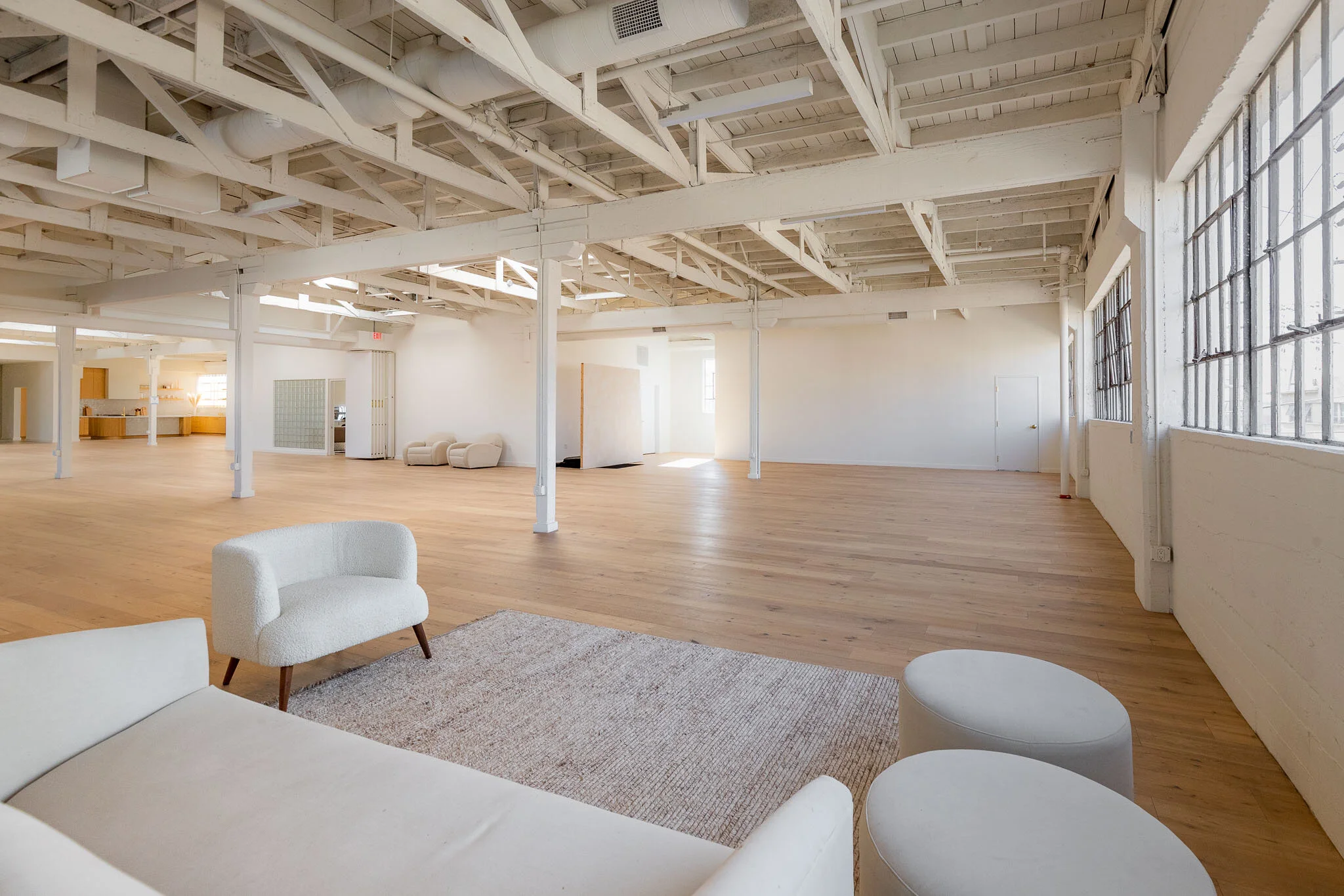 Spacious, empty loft with wooden floors, white walls, exposed ceiling beams, large windows, and minimal furniture including a white sofa, a textured armchair, and two round ottomans.