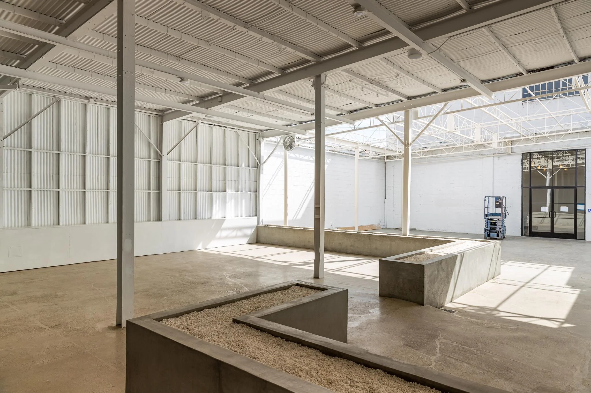 Empty indoor space with concrete planters, metal roof, white walls, and a blue scissor lift near glass doors.