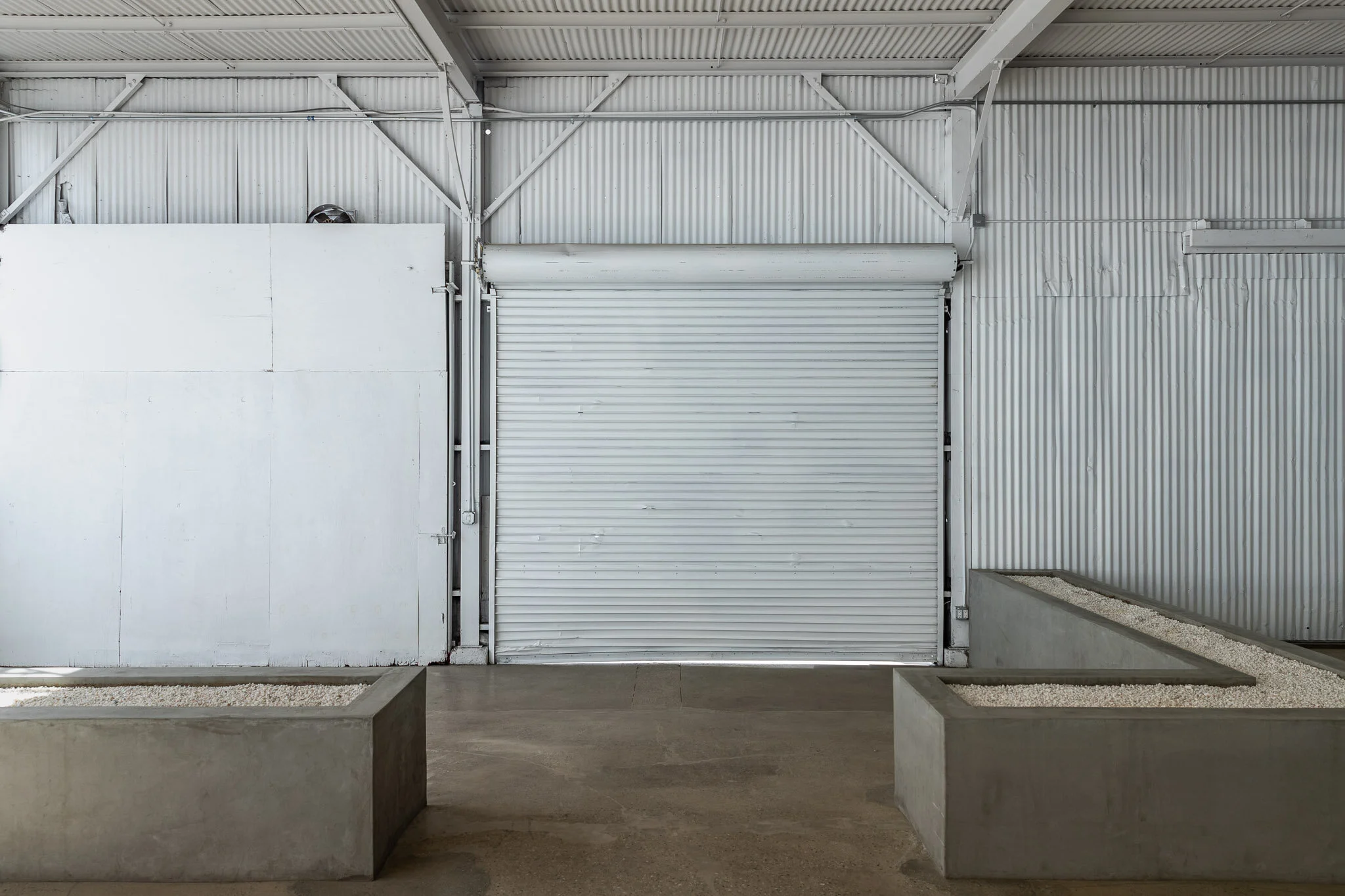 Indoor warehouse with a large metal roll-up door, concrete planters, and a corrugated metal wall.