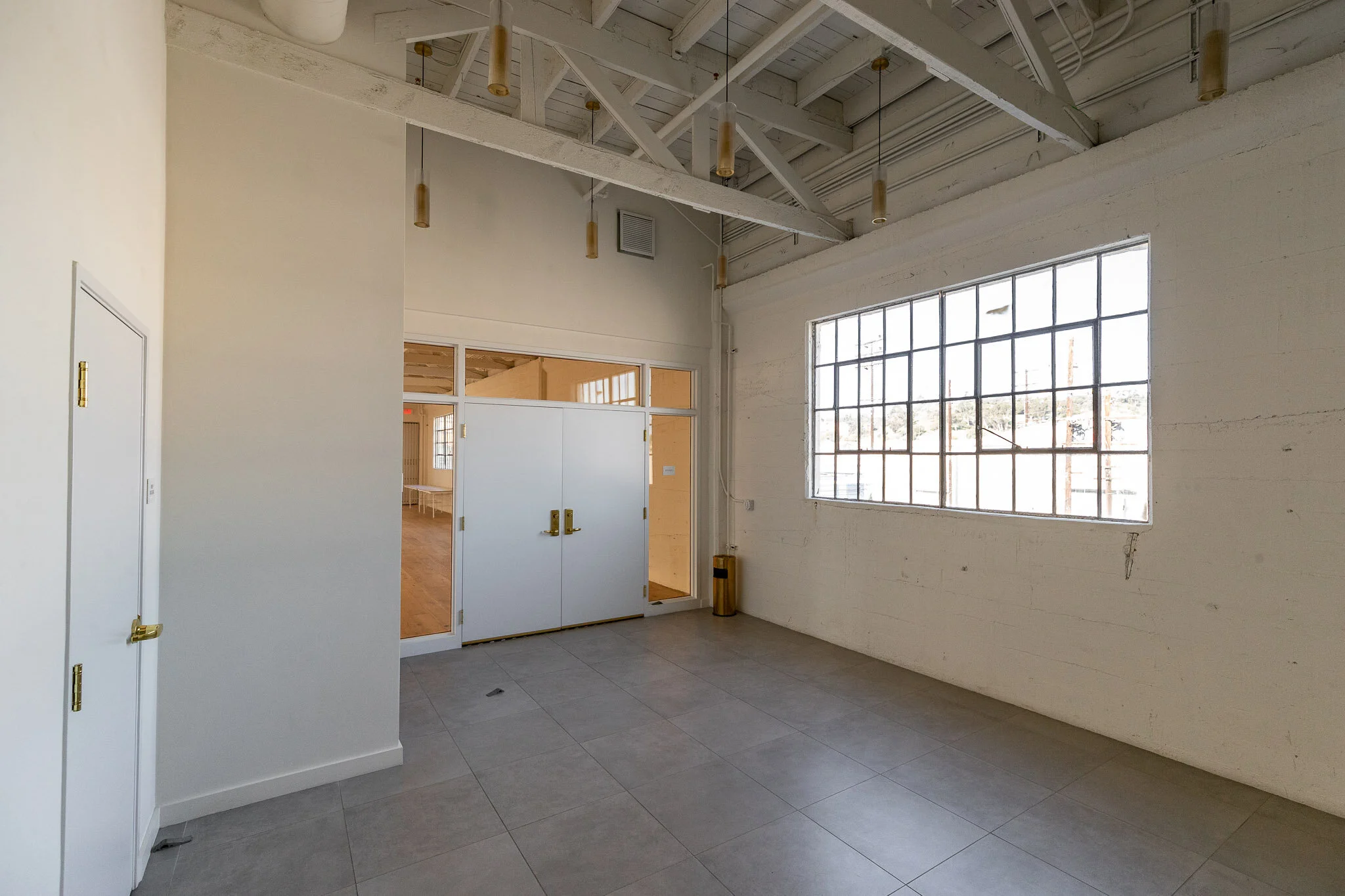 Empty room with large window, white walls, tiled gray floor, and exposed ceiling beams with hanging light fixtures.